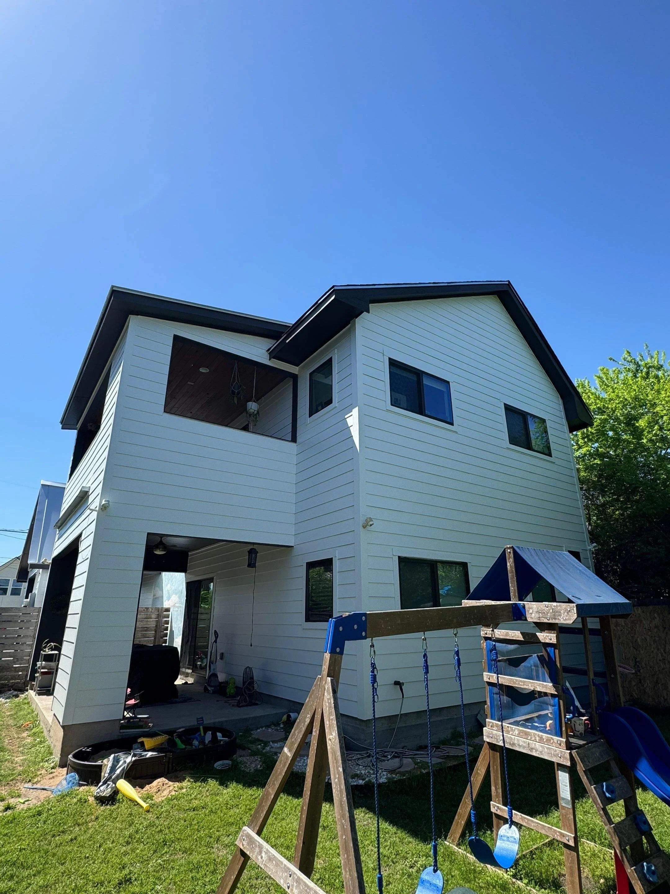 The back of a white, two-story house under renovation with a small backyard. There is a blue sky with no clouds, a wooden children's playset with swings and a slide, and some tools and equipment on the ground.