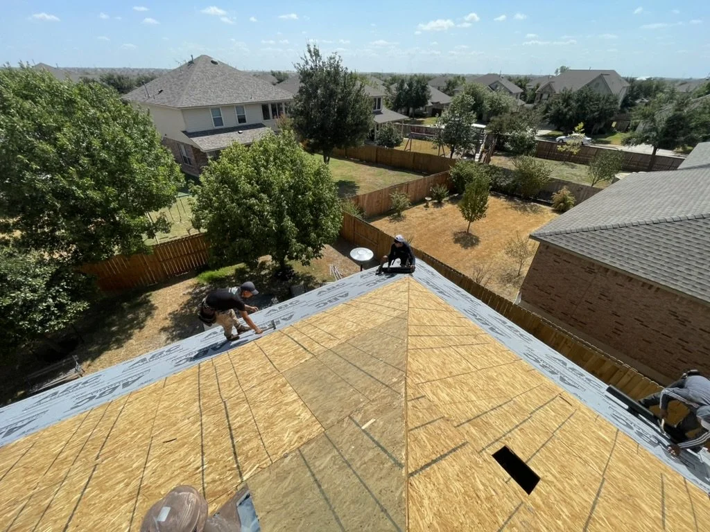 Three workers installing roofing on a house, with a backyard and neighboring houses visible in the background.