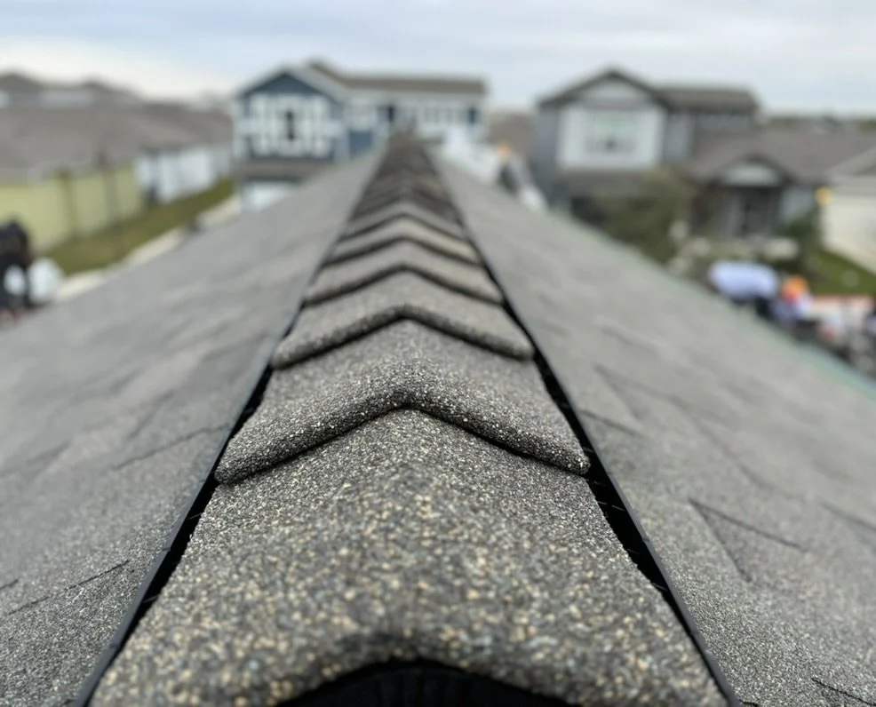Close-up of a roof with asphalt shingles, looking down the ridge toward houses in the background.