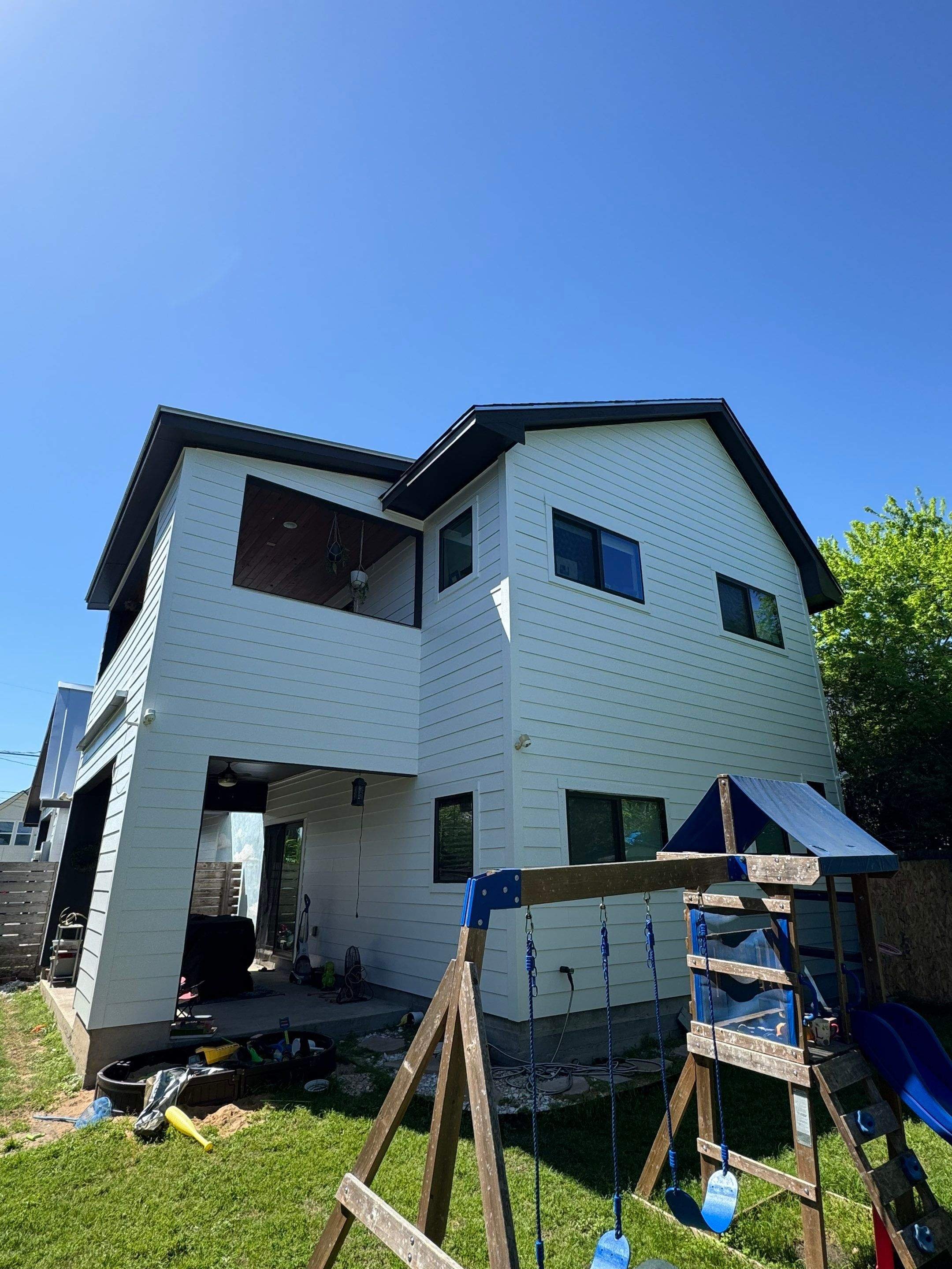 The back of a white, two-story house under renovation with a small backyard. There is a blue sky with no clouds, a wooden children's playset with swings and a slide, and some tools and equipment on the ground.