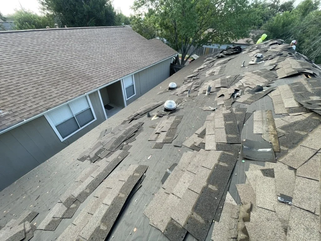 A residential roof with damaged and missing shingles, showing a steep slope and several vents. The house has a neighboring home with intact shingles and a window visible. There are workers in the background repairing the roof.