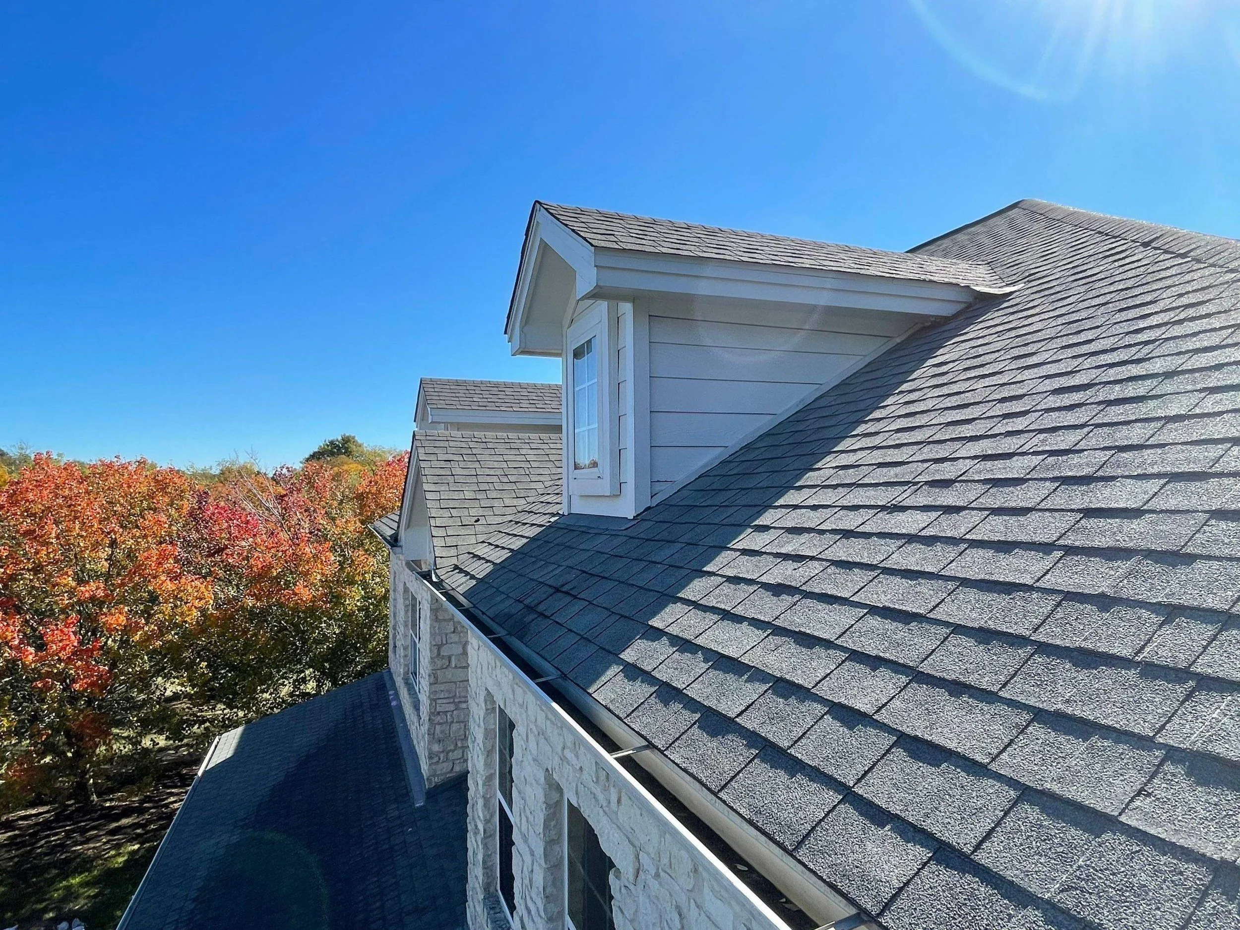 A view of a house roof with gray shingles, dormer window, and a clear blue sky in the background, with autumn-colored trees.