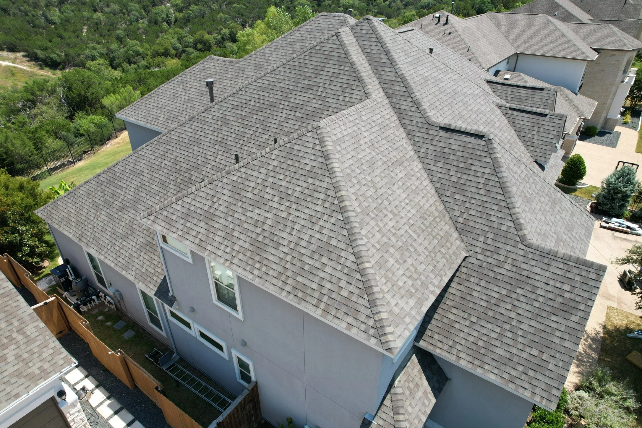 Aerial view of a multi-story house with a complex shingle roof, surrounded by trees and greenery.