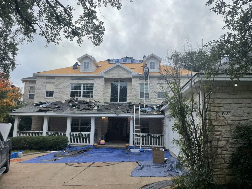 House under renovation with roof shingles removed, workers on the roof, contractor working, ladders, construction materials, and tarps laid out on the driveway.