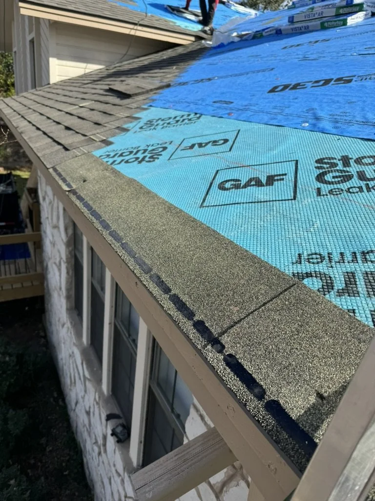 Close-up of a roof under construction with blue waterproof underlayment and asphalt shingles partially installed.