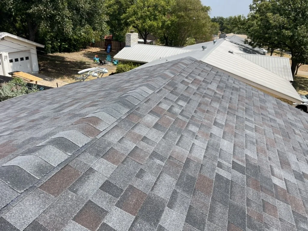 View of multiple rooftops with asphalt shingles in various shades of gray and brown, and a backyard with trees, outdoor furniture, and a small shed.