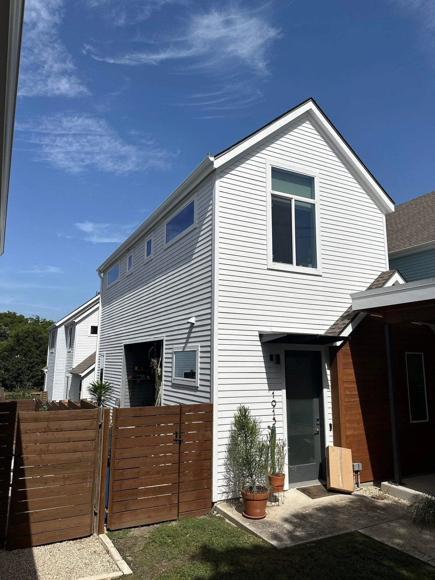 White modern house with multiple windows, a front door, potted plants, wooden fence, and neatly manicured lawn under a blue sky.