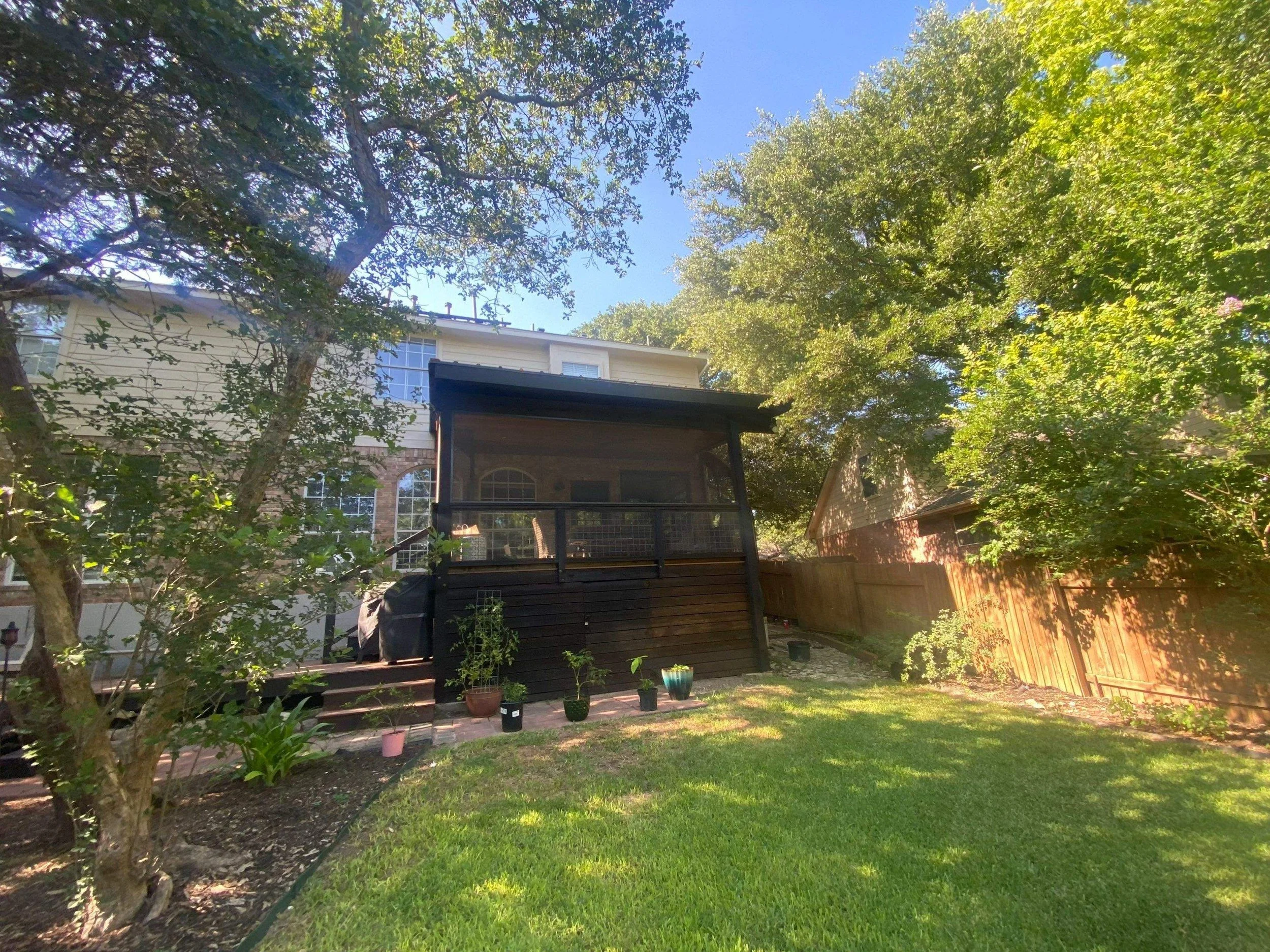 Backyard with grass lawn, potted plants on steps, trees, and a black screened porch attached to a two-story house.