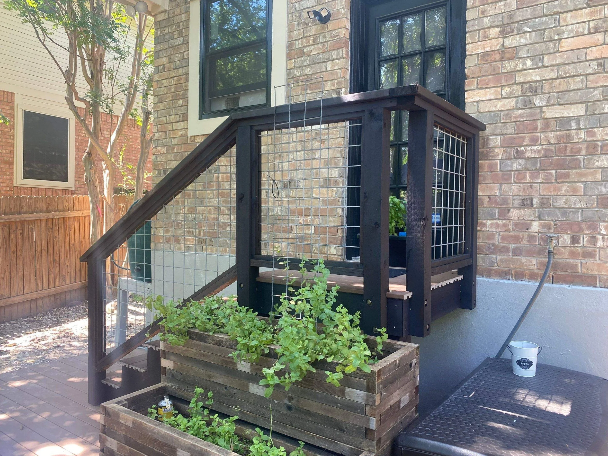 Backyard porch with a small black wooden deck and stairs, with planter boxes filled with green plants, a small white bucket on a black table, and a brick house wall with windows surrounding the porch.