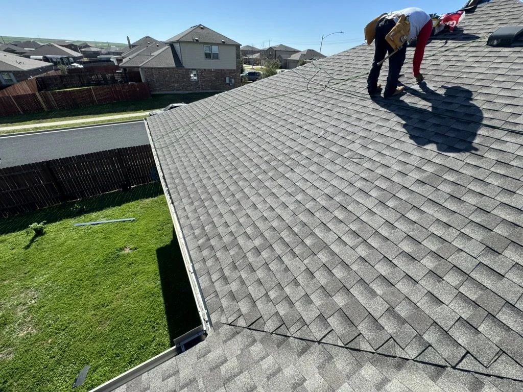 A worker on a gray shingled roof using a tool, with a cloudy sky and residential neighborhood in the background.