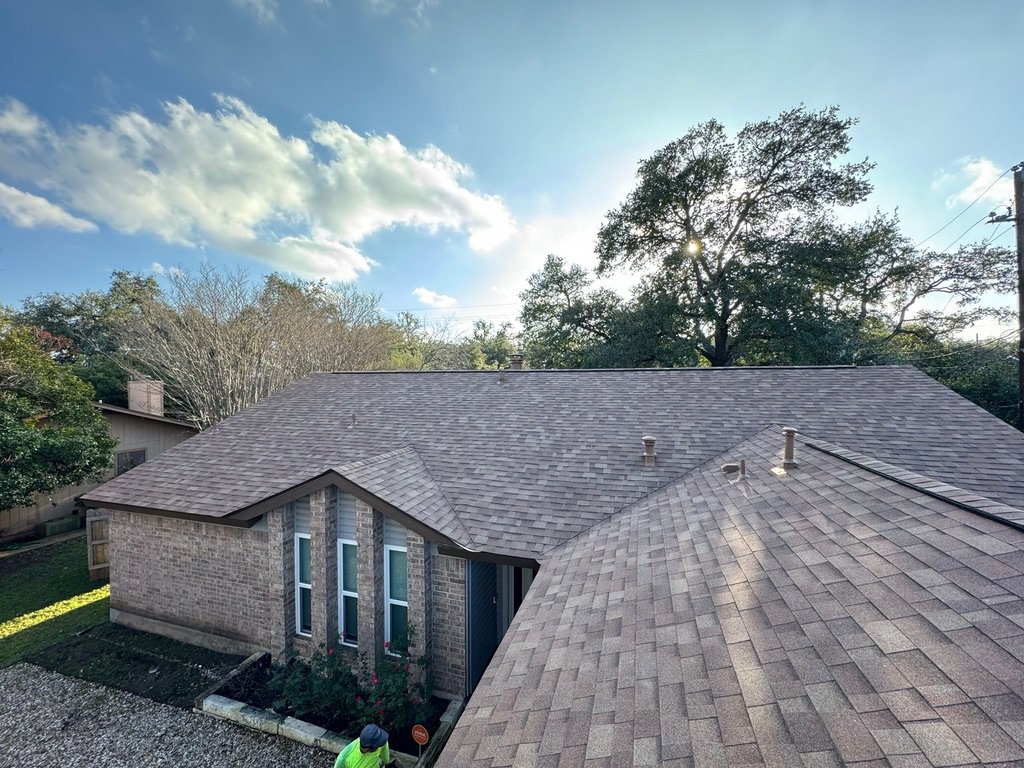 A residential house with a shingled roof, brick exterior, and a small front yard with plants. The photo is taken outdoors during the daytime with a partly cloudy sky.