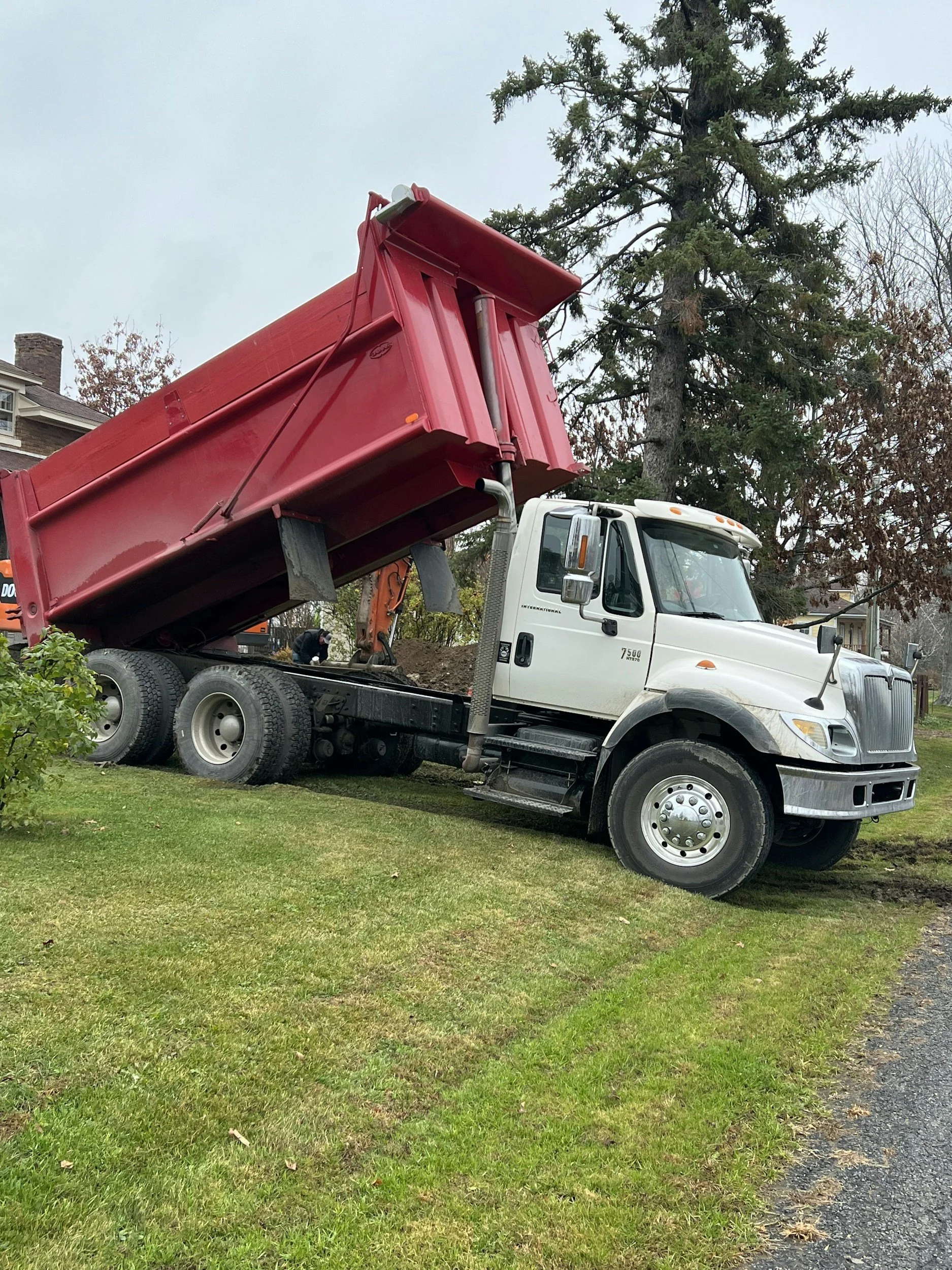 A white dump truck with a red bed raised, positioned on a lawn with trees in the background.