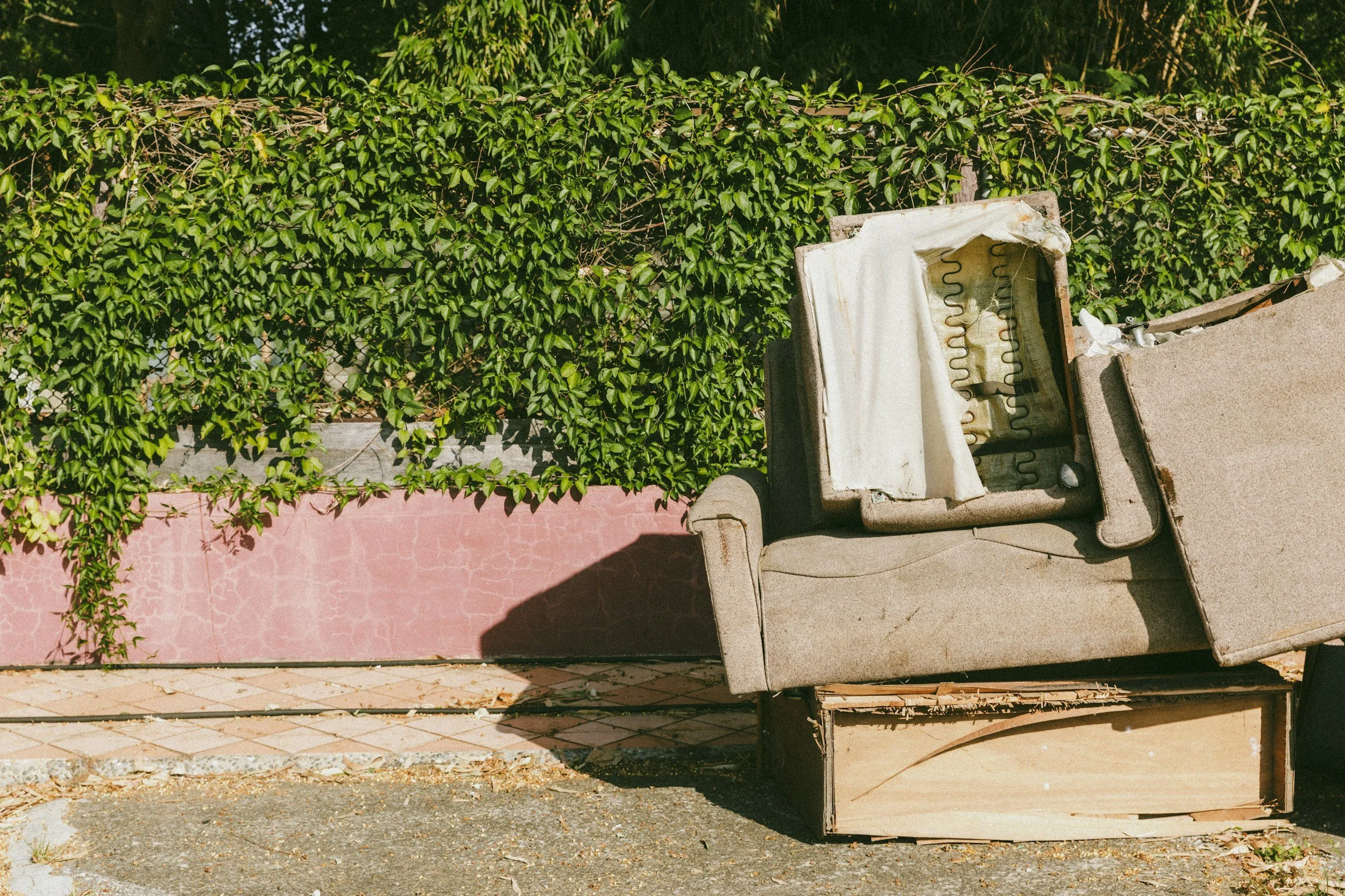 A worn-out beige armchair with a white, torn cushion and exposed springs, placed outdoors against a green hedge and pink wall.