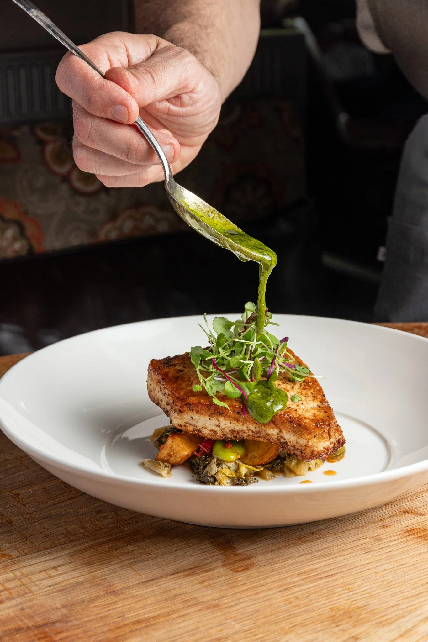 A chef is garnishing a plated dish with green sauce and microgreens. The dish features a seared piece of fish on a bed of vegetables, all on a white plate.