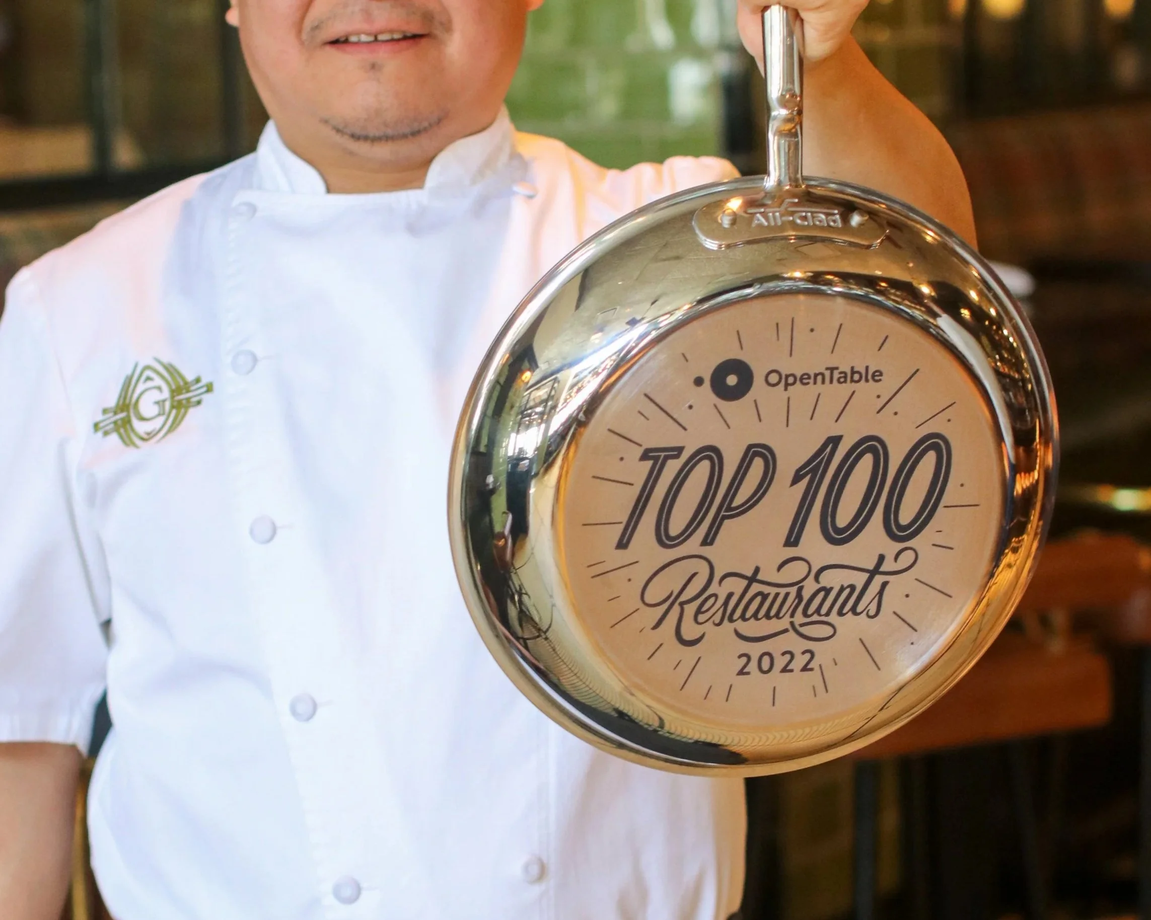 Close-up of a chef holding a shiny round clock with the words "Top 100" and "Restaurant" and the year "2022" on its face. The chef is smiling and wearing a white chef's coat with a green embroidery on the sleeve.