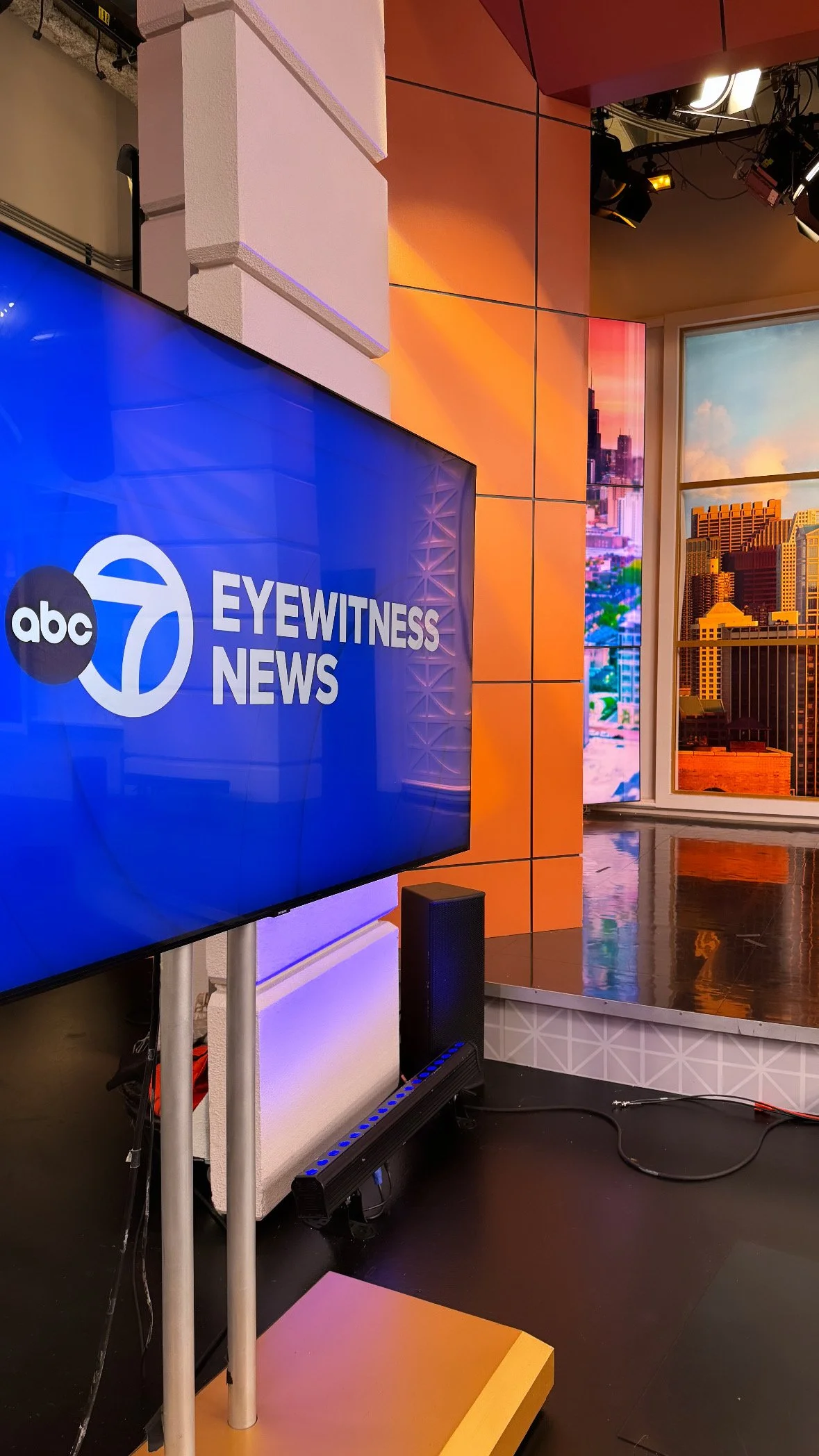 Television screen displaying the logos of ABC and Eyewitness News in a studio with orange and beige walls, large windows showing city buildings, and studio lights hanging from the ceiling.