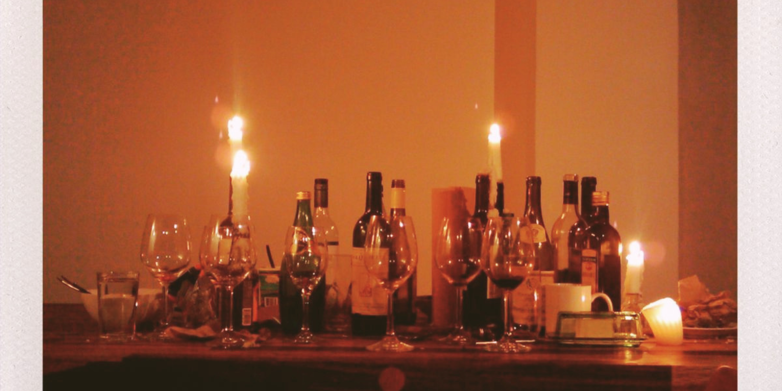 A table with various bottles of alcohol, empty wine glasses, and lit candles creating a warm ambiance.