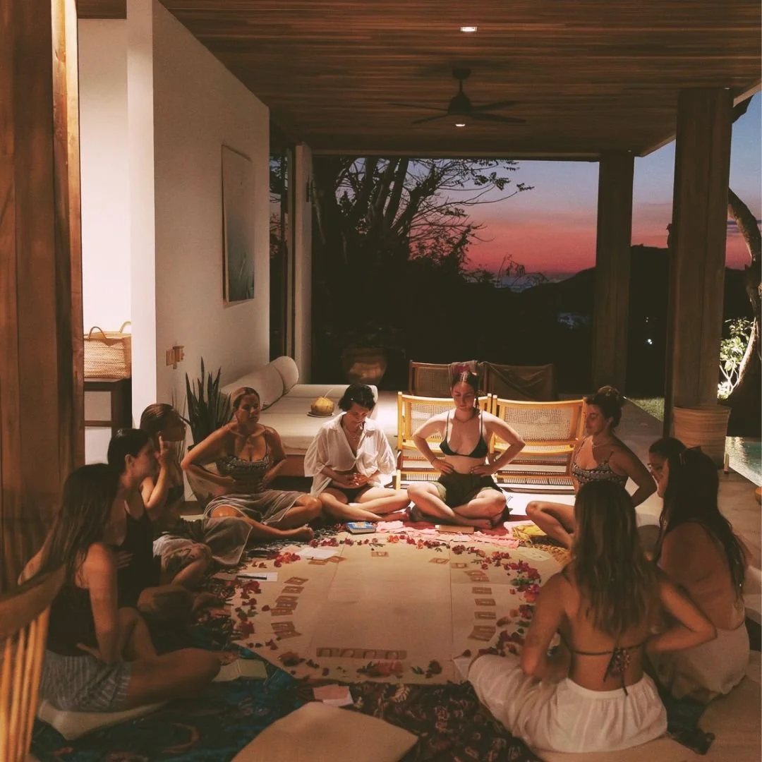 Group of women sitting in a circle on a blanket. The setting is a covered outdoor patio with a sunset view.