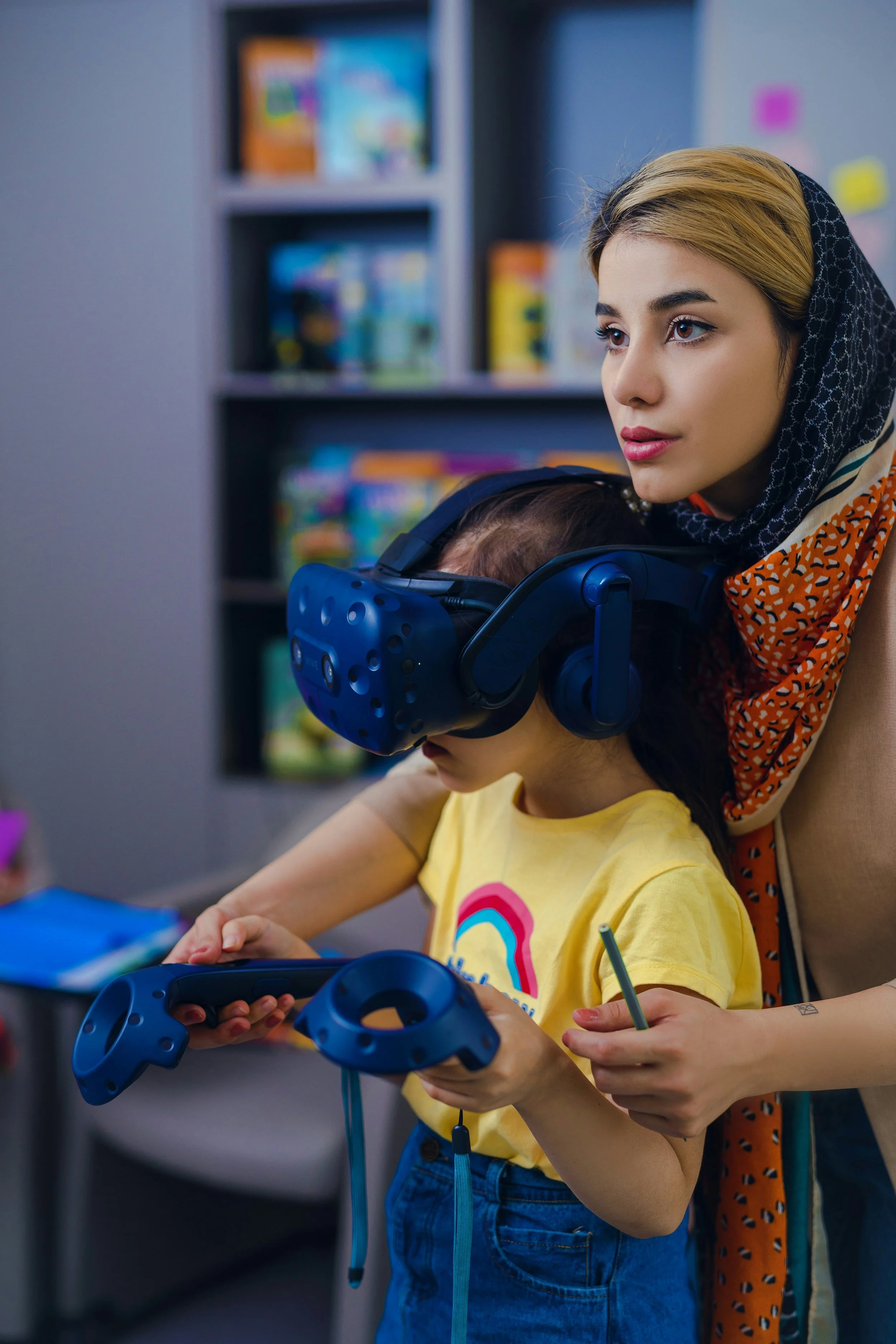 A woman with blonde hair, wearing a black and white patterned headscarf and an orange patterned dress, is helping a young girl with dark hair and a yellow t-shirt to use a virtual reality headset. The girl is holding a game controller, and the setting appears to be a room with bookshelves in the background.