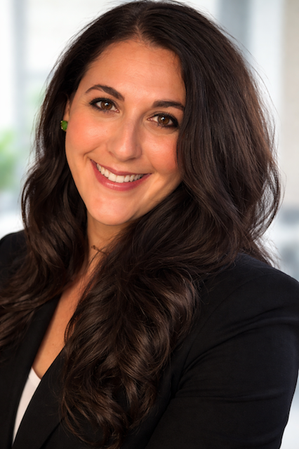 Close-up of a smiling woman with long, dark, wavy hair, wearing a black blazer and green earrings, in a professional indoor setting.