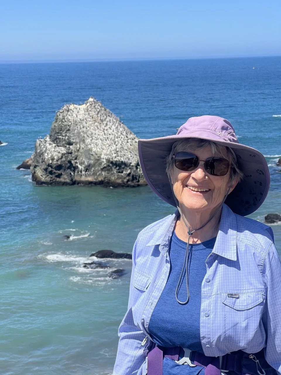Smiling woman in sunglasses and a wide-brimmed hat standing near the ocean with a large rock in the background.