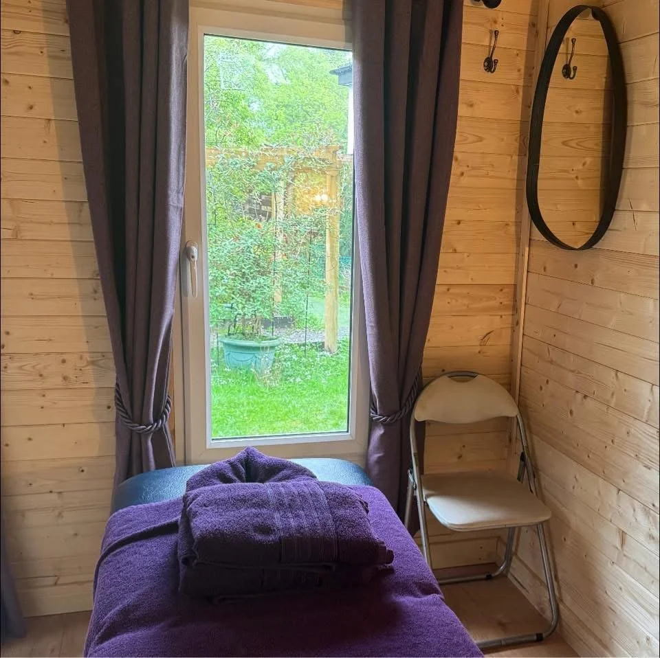 Massage table in a log cabin in my garden with purple towels and a view from the window on to a honeysuckle plant.