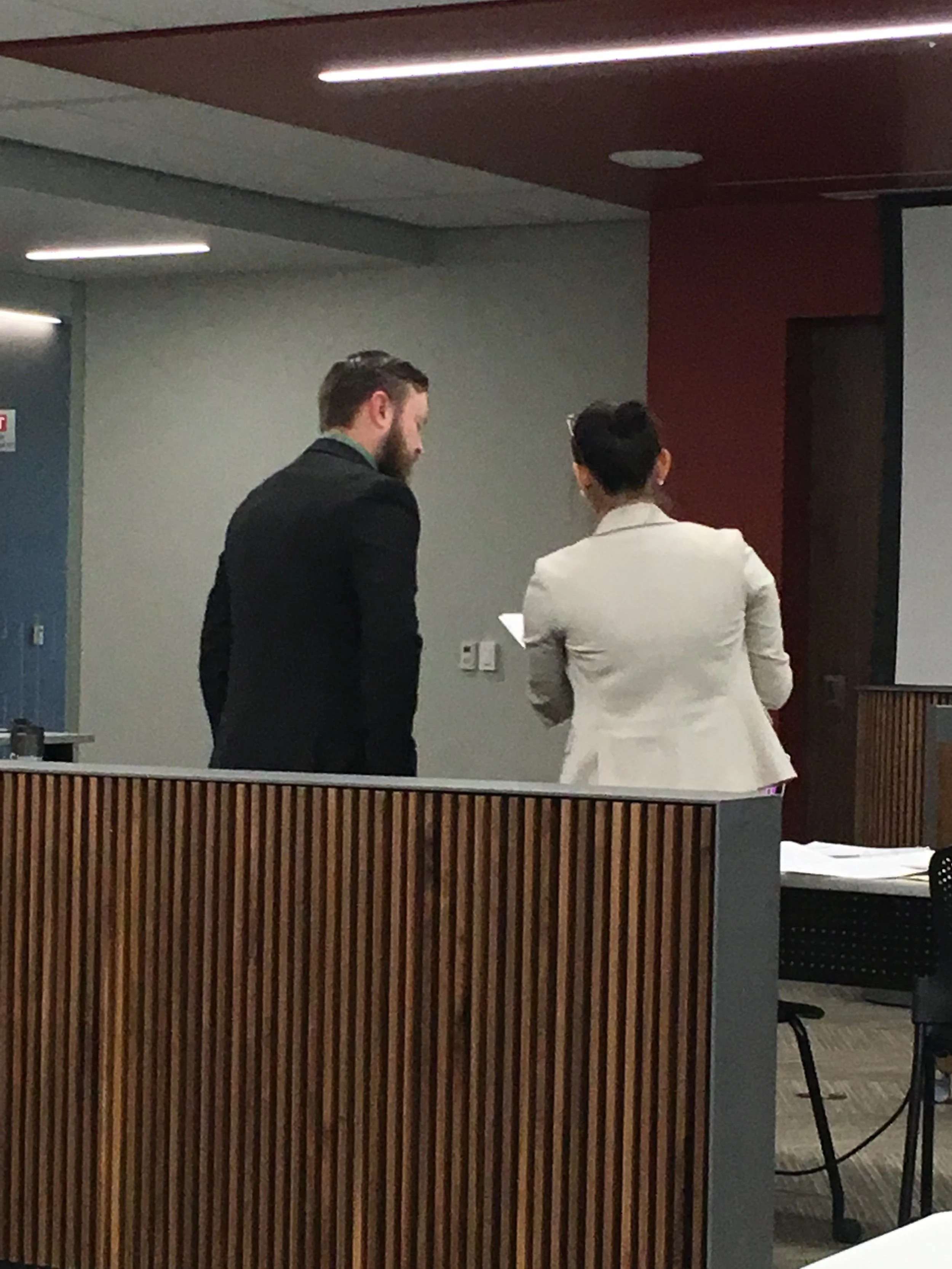 Two people, one male and one female, are engaged in conversation in a modern indoor setting with gray walls and wood accents.