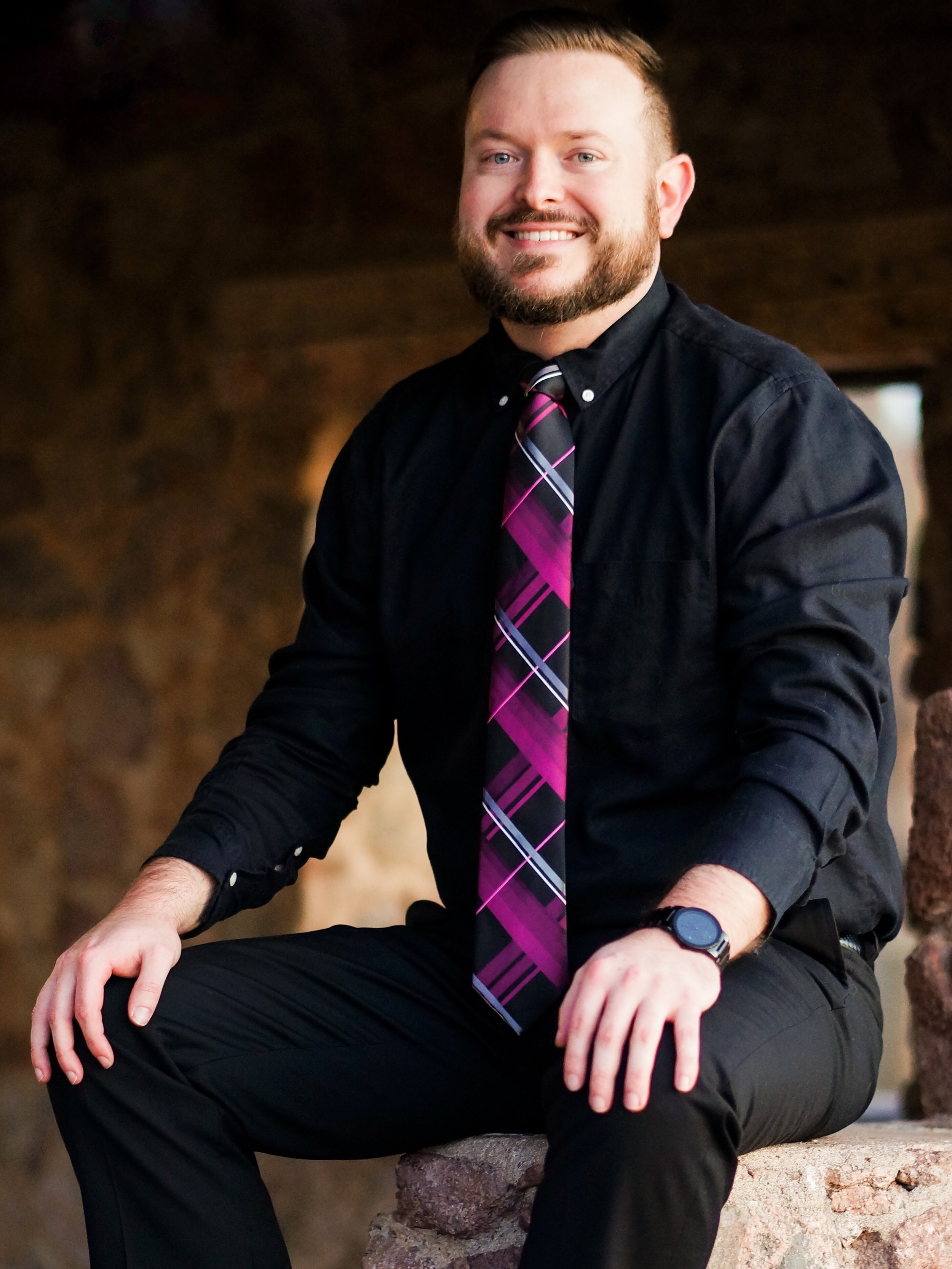 A man with a beard and short hair, smiling and dressed in a black shirt and black pants, sitting on a stone ledge in a dimly lit environment.