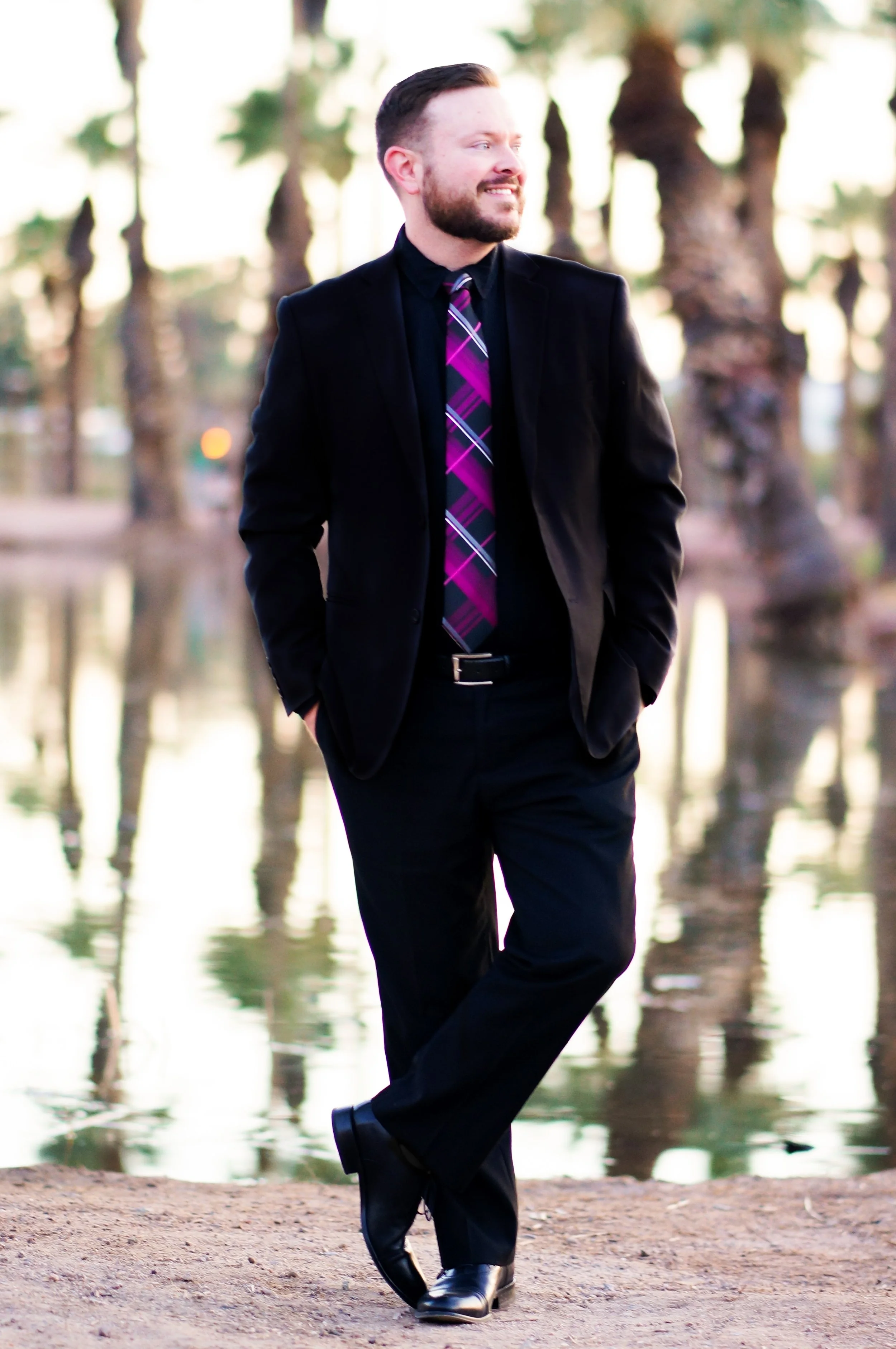 A man dressed in a black suit with a purple, pink, and white plaid tie standing near water with trees in the background during sunset.