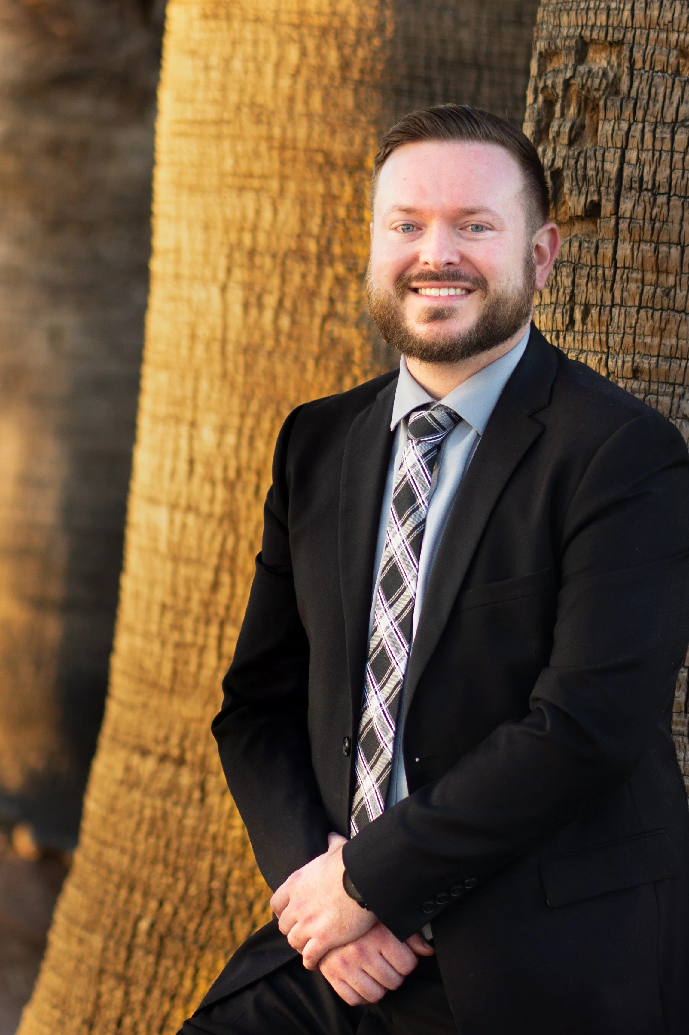 A man with a beard and short hair wearing a business suit, striped tie, and light blue shirt, smiling and sitting with his hands clasped in front of a large wooden texture background.