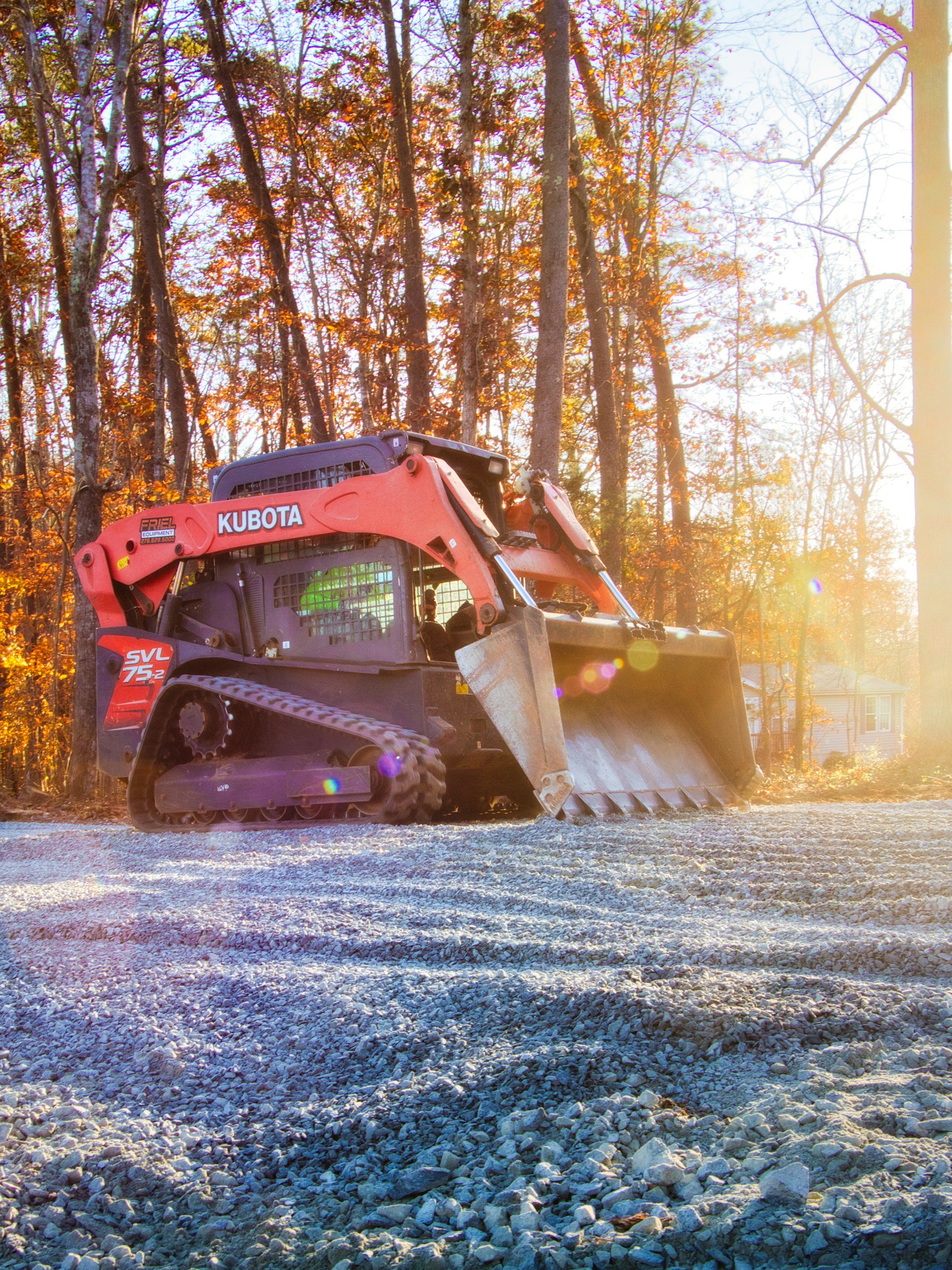 A construction vehicle, a Kubota compact excavator, is working on a gravel surface in a wooded area during sunset, with autumn leaves on the trees.