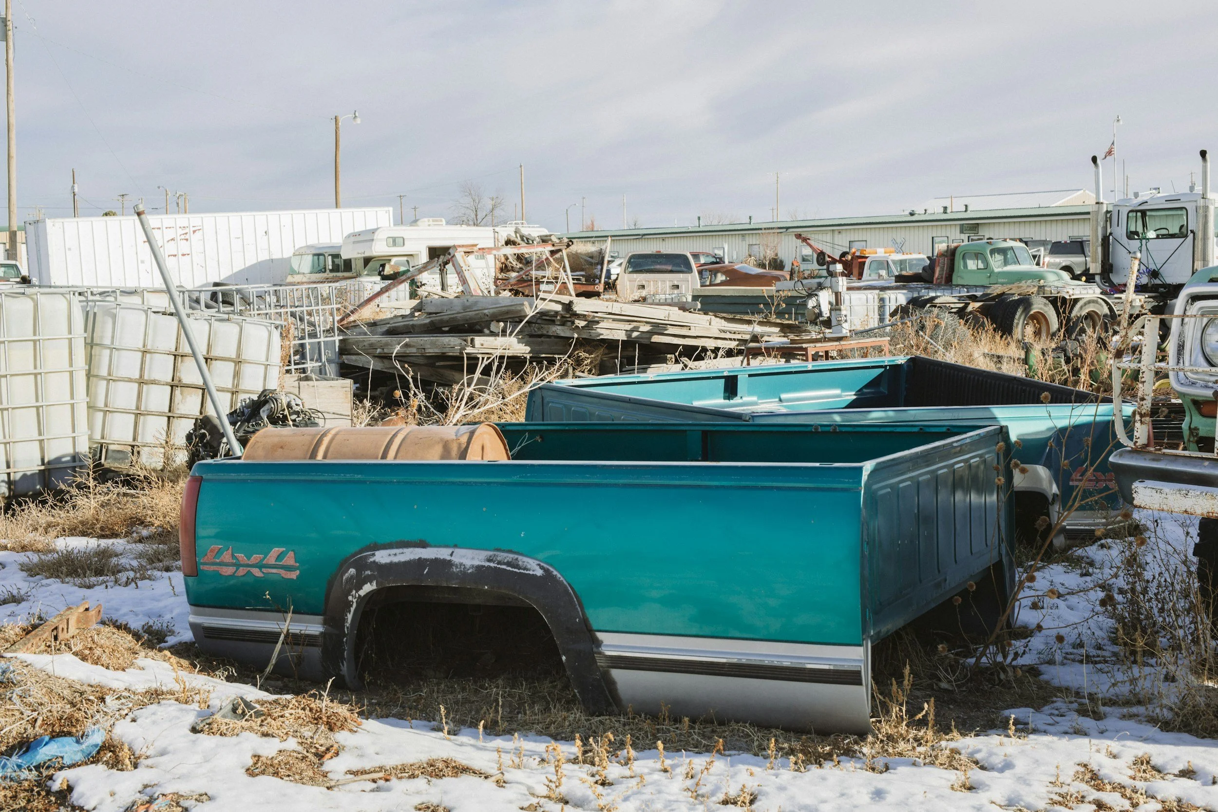 A junkyard filled with old, rusted vehicles, trailers, and debris, with some snow on the ground and a partly cloudy sky overhead.