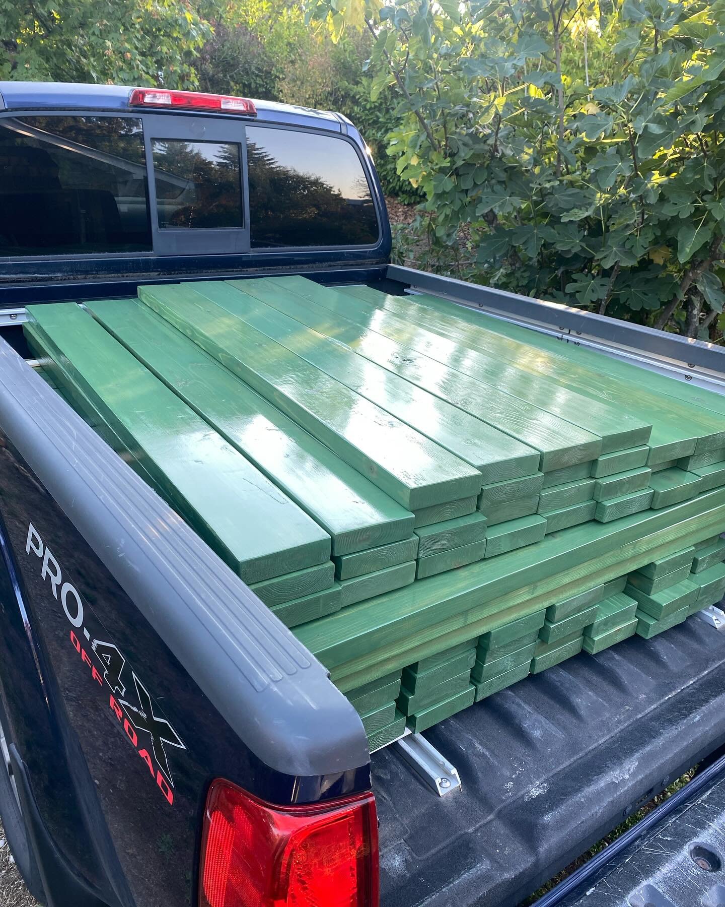 Pickup truck carrying a load of green wooden planks in its truck bed, parked outdoors with trees and bushes in the background.