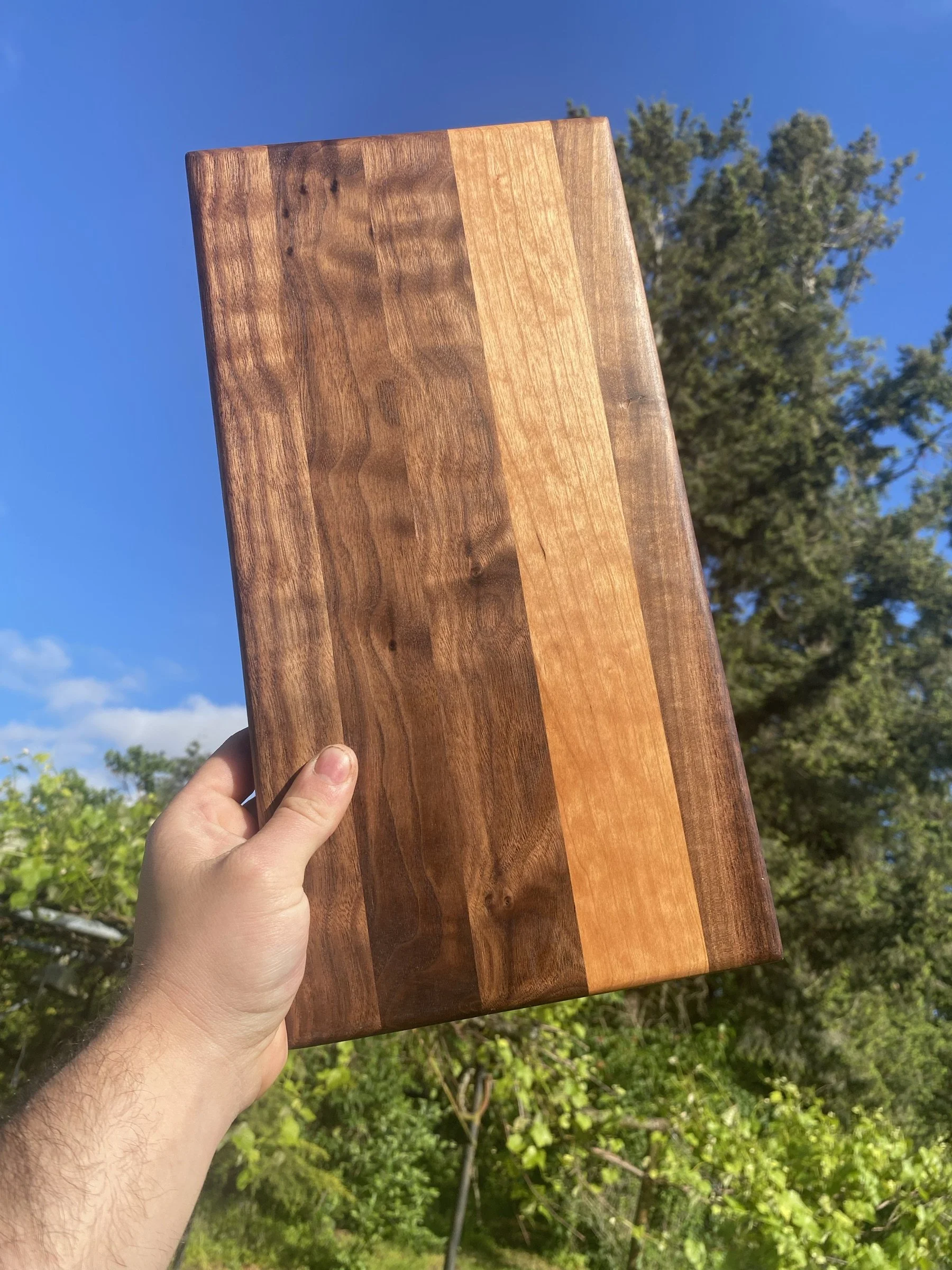A walnut and cherry cutting board held up against a blue sky.
