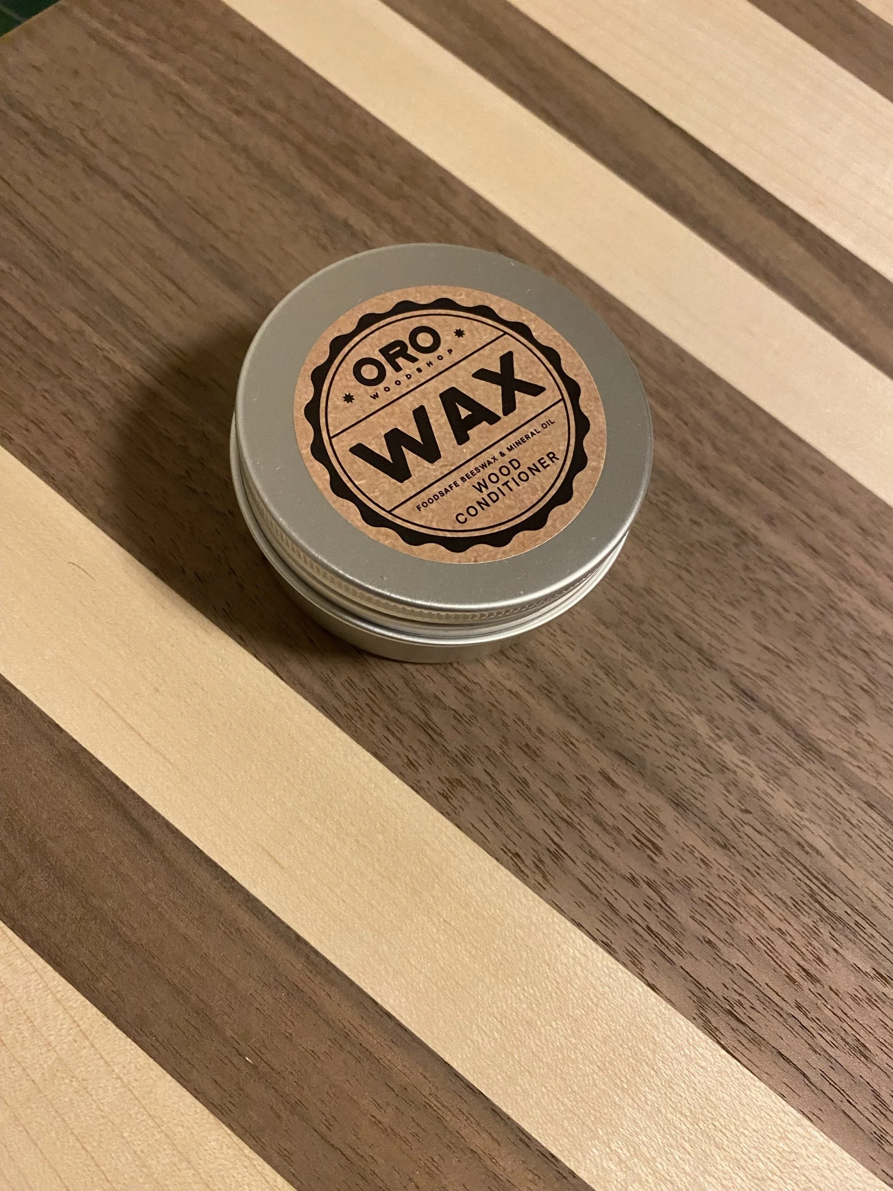 A tin of Oro's own board wax resting on a striped hardwood cutting board.