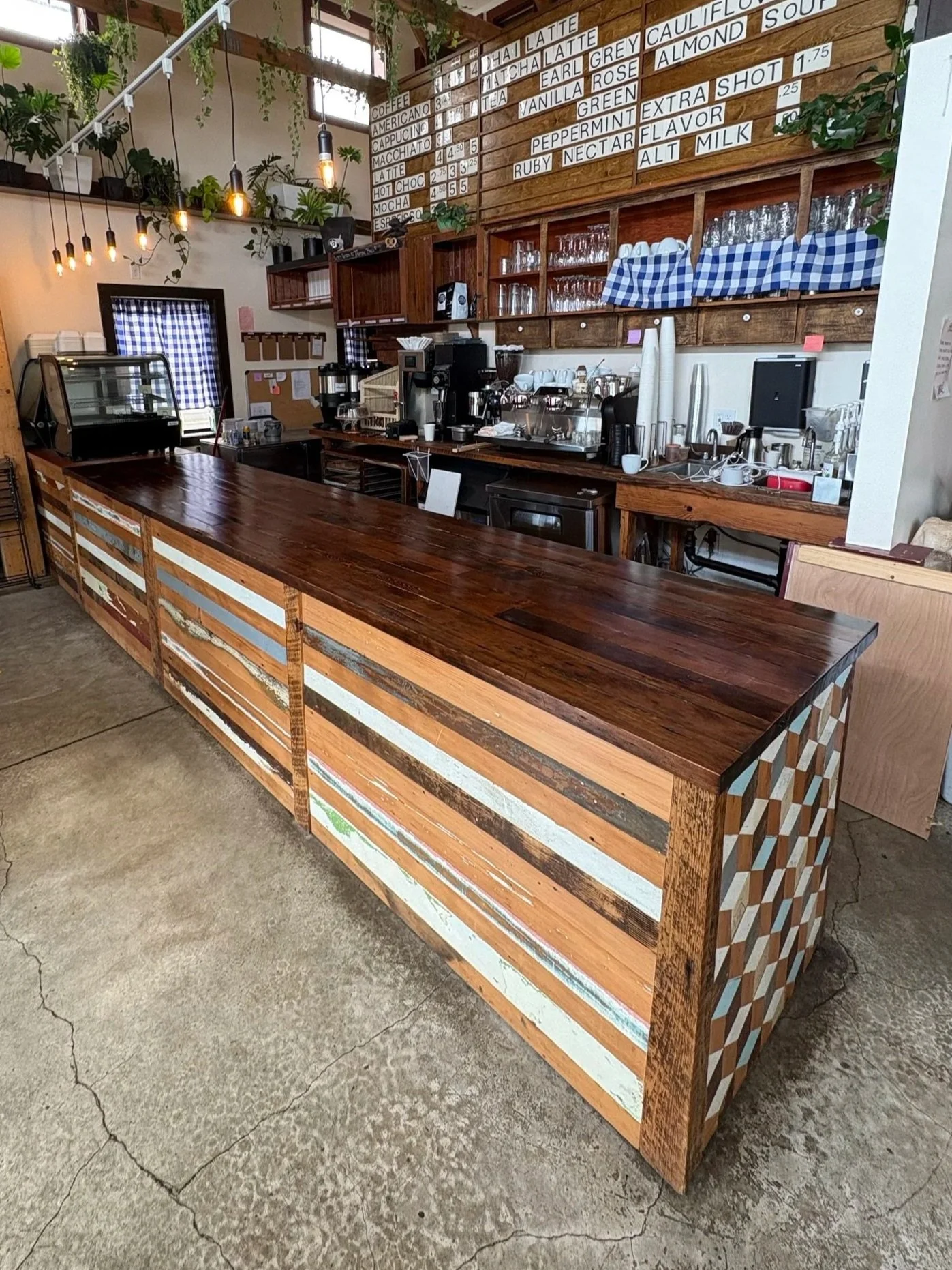 Interior of a bakery with a wooden counter, hanging lights, wall menu, coffee machines, glassware, and a window with checkered curtains.