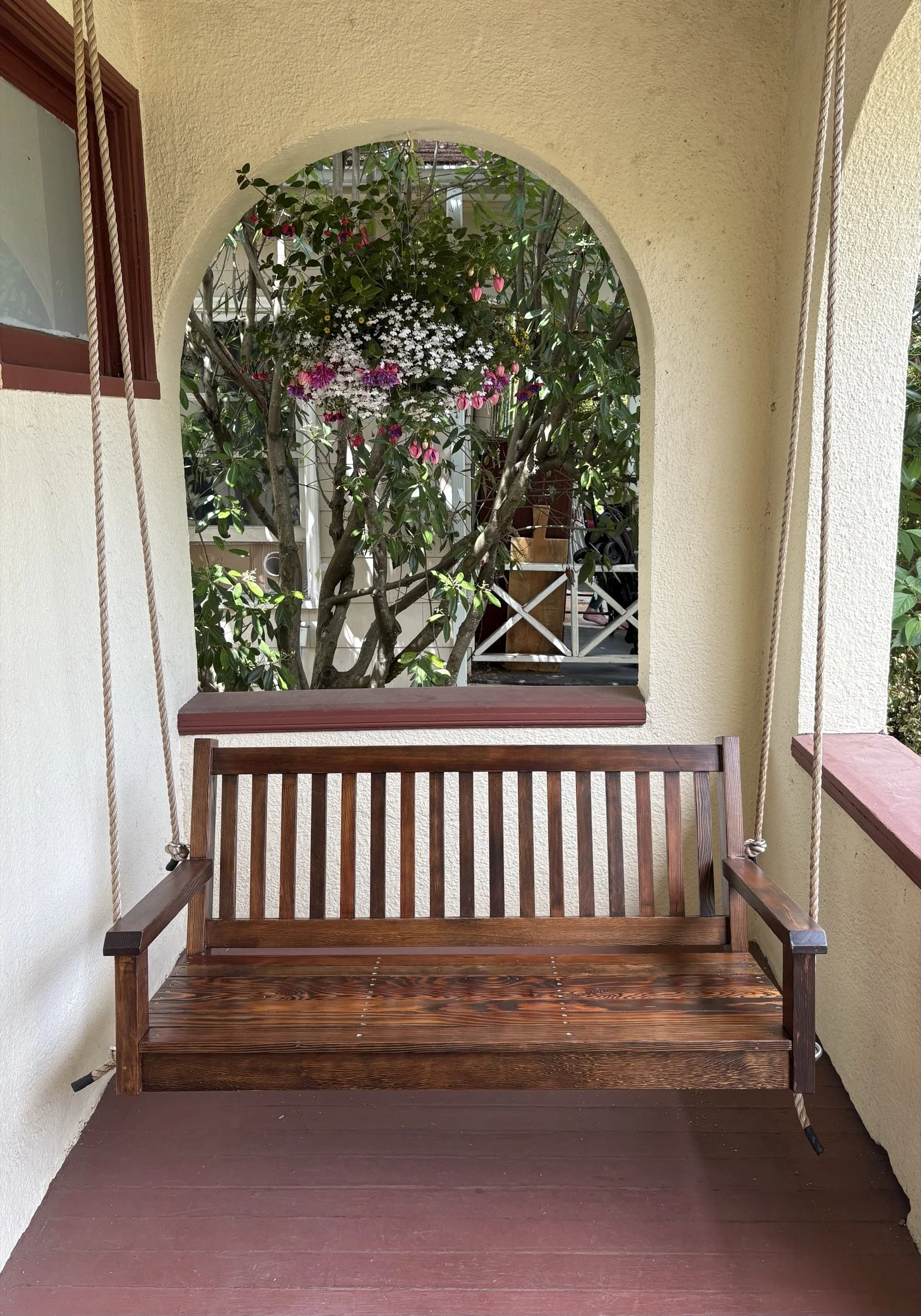 Wooden porch swing with a backrest and armrests, hanging in front of beige walls with a window and trees visible through an arch opening behind.