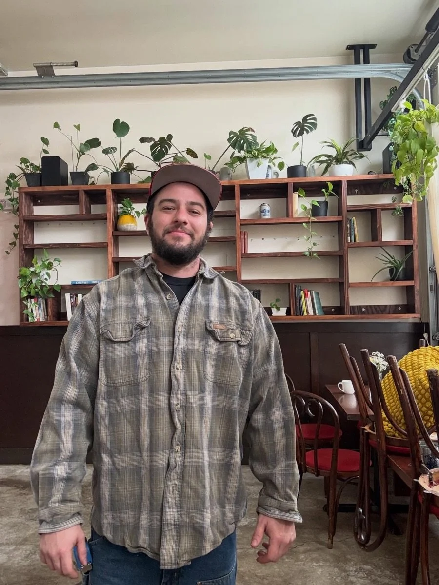 A person with a beard wearing a baseball cap and plaid shirt standing inside a room with a bookshelf and plants in the background.
