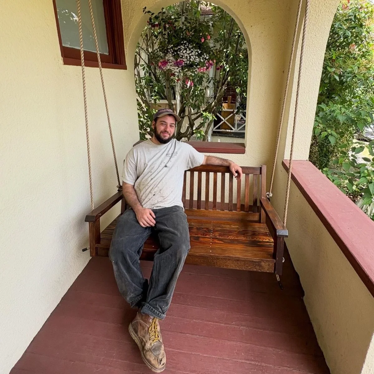 A person with a beard sitting on a wooden porch swing, wearing a white T-shirt, dark jeans, and yellow work boots, with one arm resting on the swing's backrest, surrounded by greenery and a flowering plant.