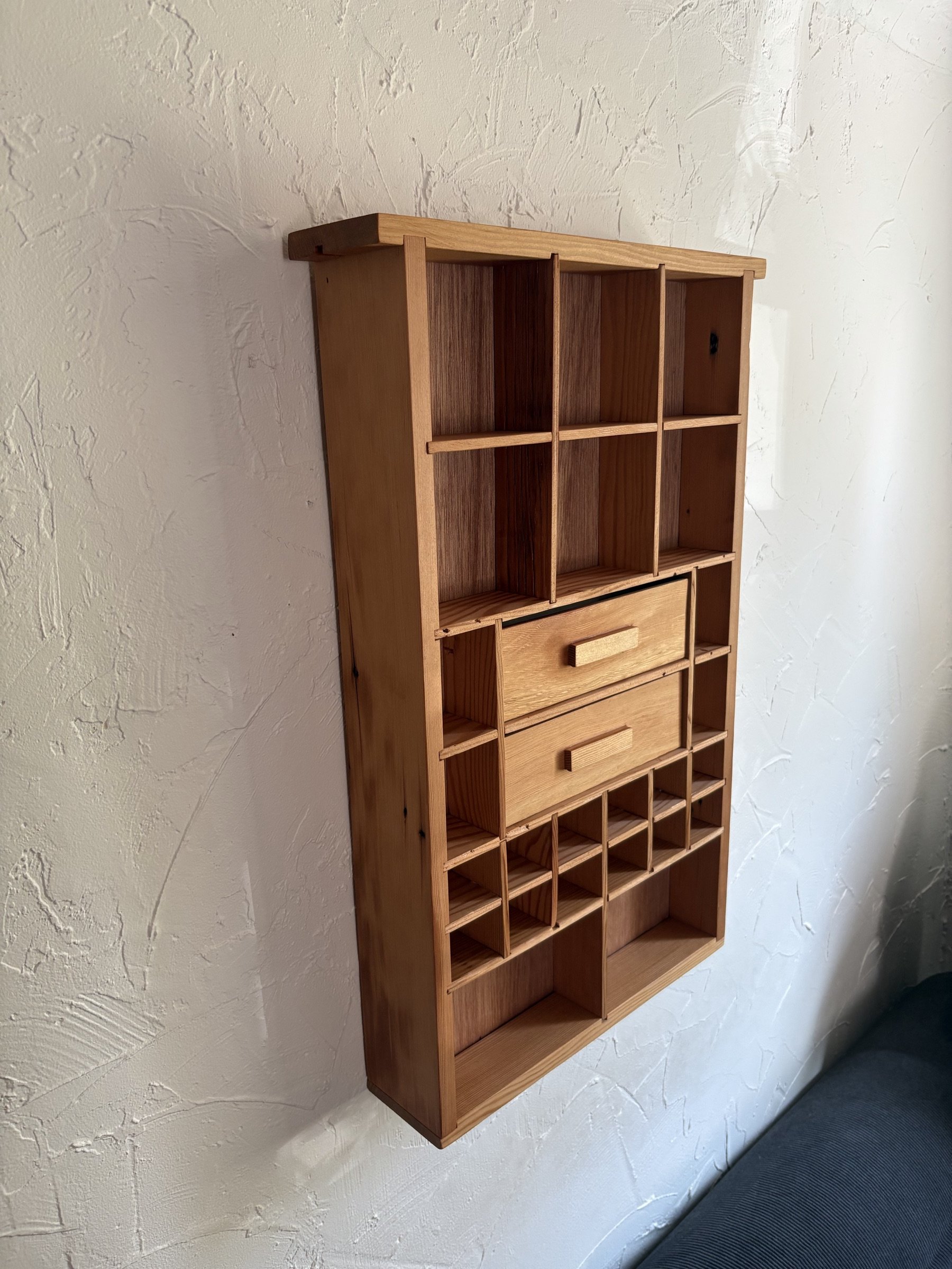 A lighter oak curio shelf with two drawers and open compartments, mounted on a plaster wall.