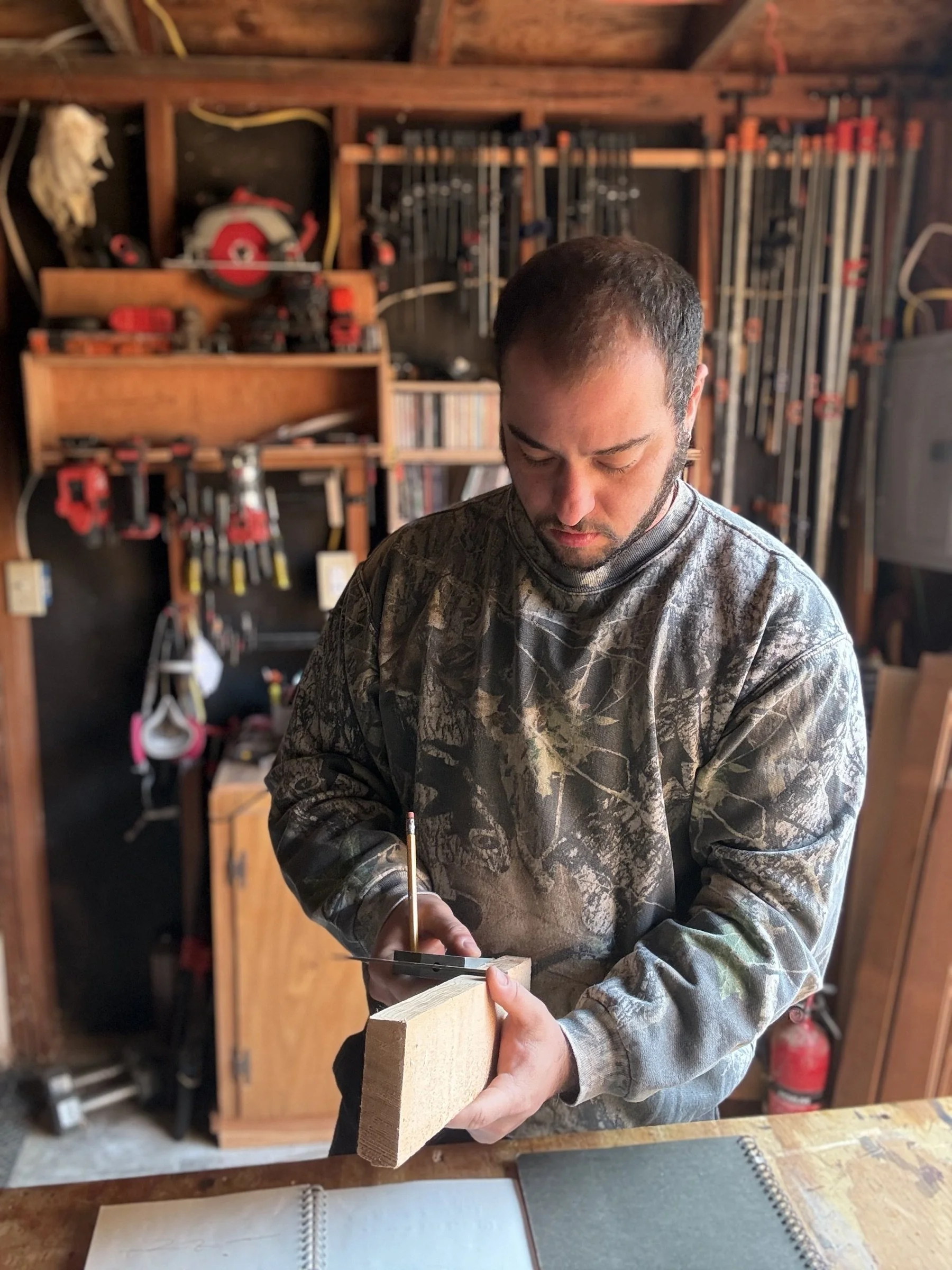 A person in camouflage shirt working in a woodworking shop, holding a piece of wood and using a tool, with shelves of clamps and tools in the background.