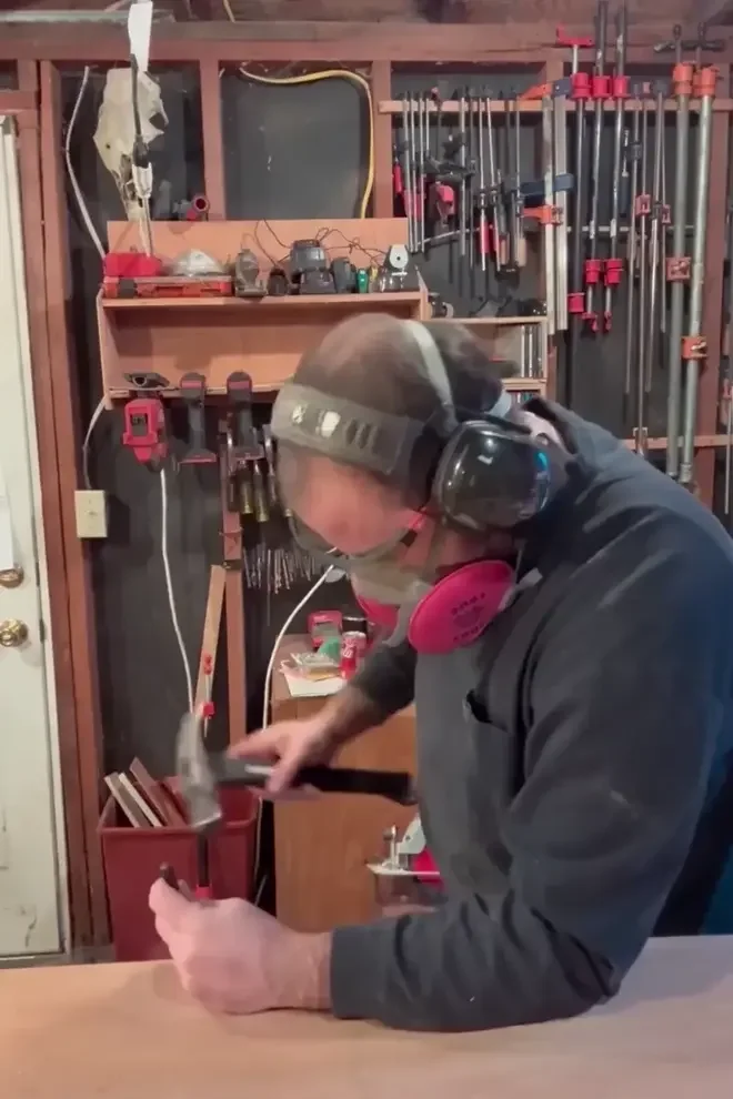 A person wearing hearing protection and a face mask is using a hammer on a piece of metal in a workshop. The background shows various clamps, tools, and equipment on shelves and hanging on the wall.