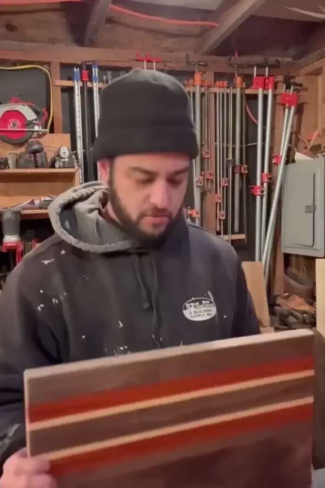 A person with a beard in a black beanie and hoodie inspecting a wooden piece with colorful stripes in a woodworking workshop, with tools and clamps on the wall behind them.
