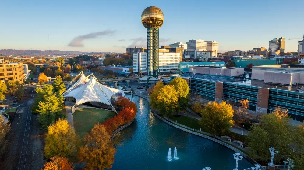 A cityscape with a park, a waterway, and a distinctive spherical tower in the background. Fall foliage is visible on trees along the water and around the park.