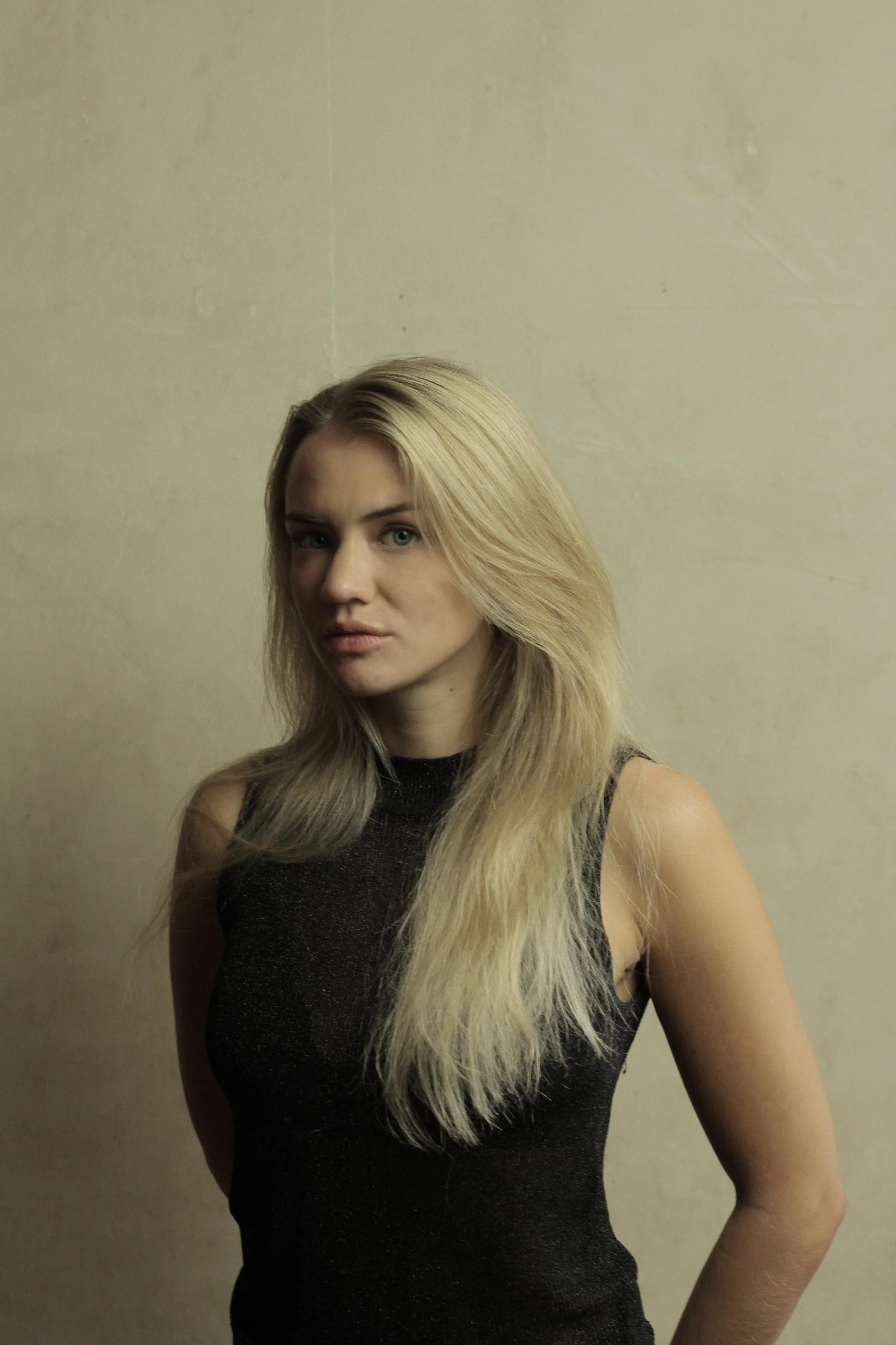 A woman with long blonde hair wearing a black sleeveless top standing against a plain beige wall.