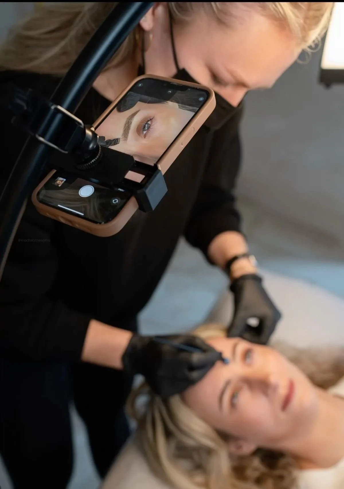 A makeup artist is applying makeup to a woman's face while taking a close-up photo with a smartphone. The phone's screen shows a zoomed-in view of the woman's eye and eyebrow.
