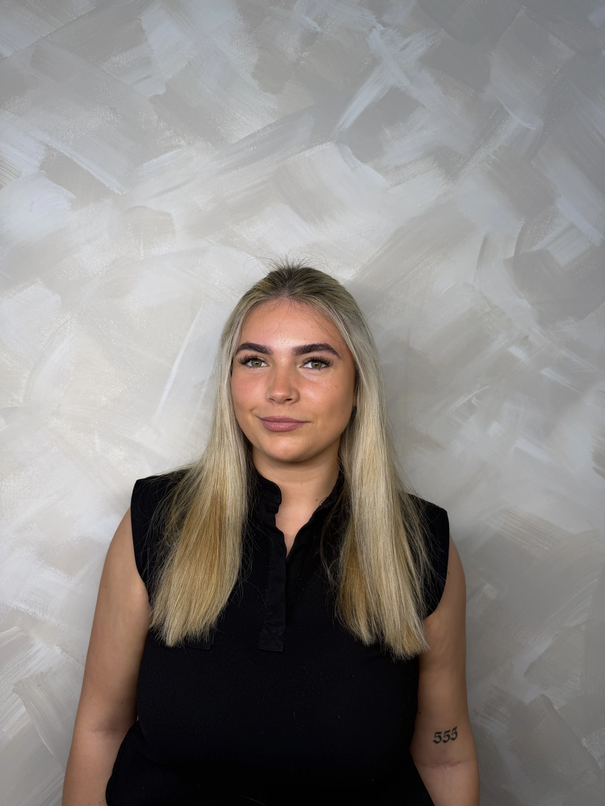 A woman with long blonde hair and green eyes is looking at the camera. She has a subtle smile and is wearing a sleeveless black shirt. Her right arm has a tattoo with the number '555'. The background is a textured wall with abstract brushstrokes in shades of gray and white.