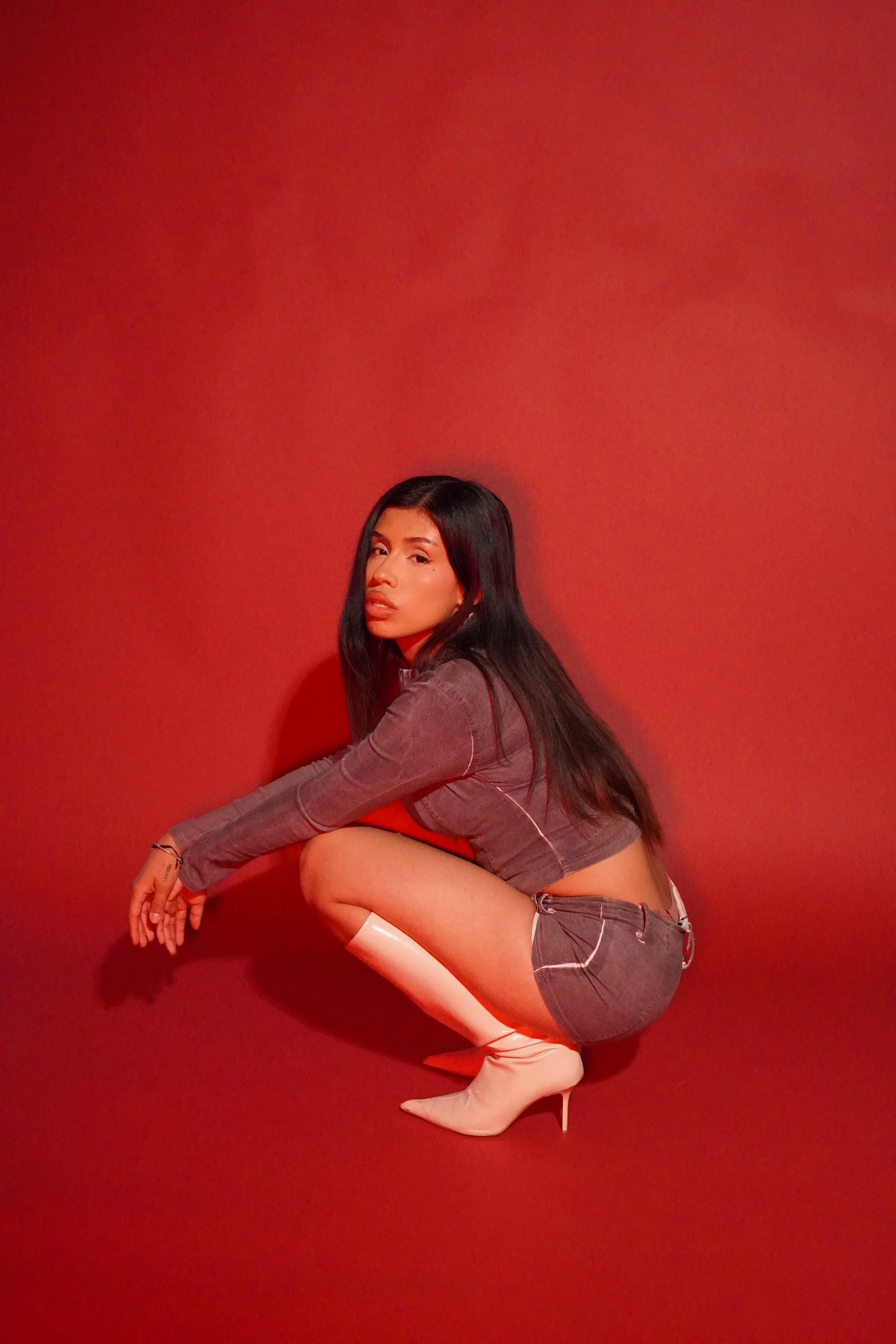 A woman with long dark hair and a confident expression, dressed in a plaid jacket and matching plaid shorts, poses against a solid red background, wearing white high-heeled boots.