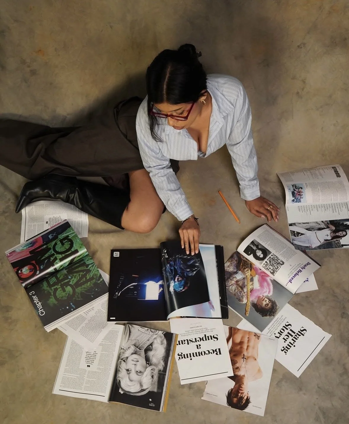 A woman sits on the floor surrounded by open magazines and newspapers, reading and browsing through them. She is wearing glasses, a pinstripe shirt, and black boots. The setting appears to be an indoor space with a concrete floor and a plain wall.