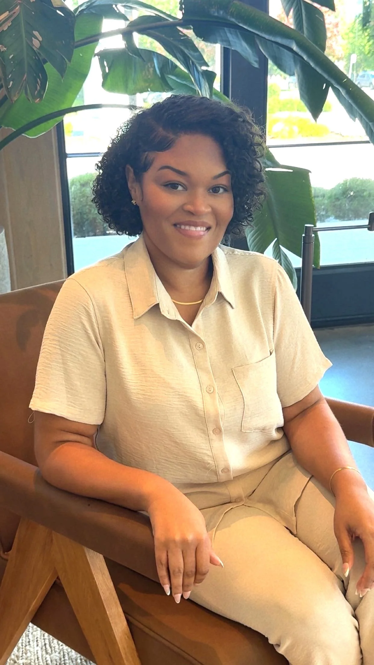A woman with curly black hair sitting in a chair in a room with large green plants and windows in the background.